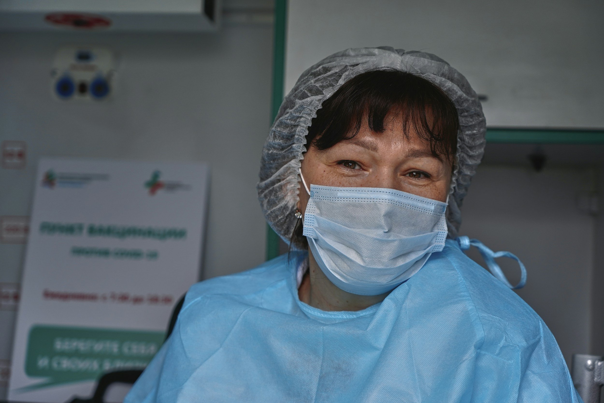 Medical professional wearing a surgical mask, hair cap, and protective gown in a healthcare setting.