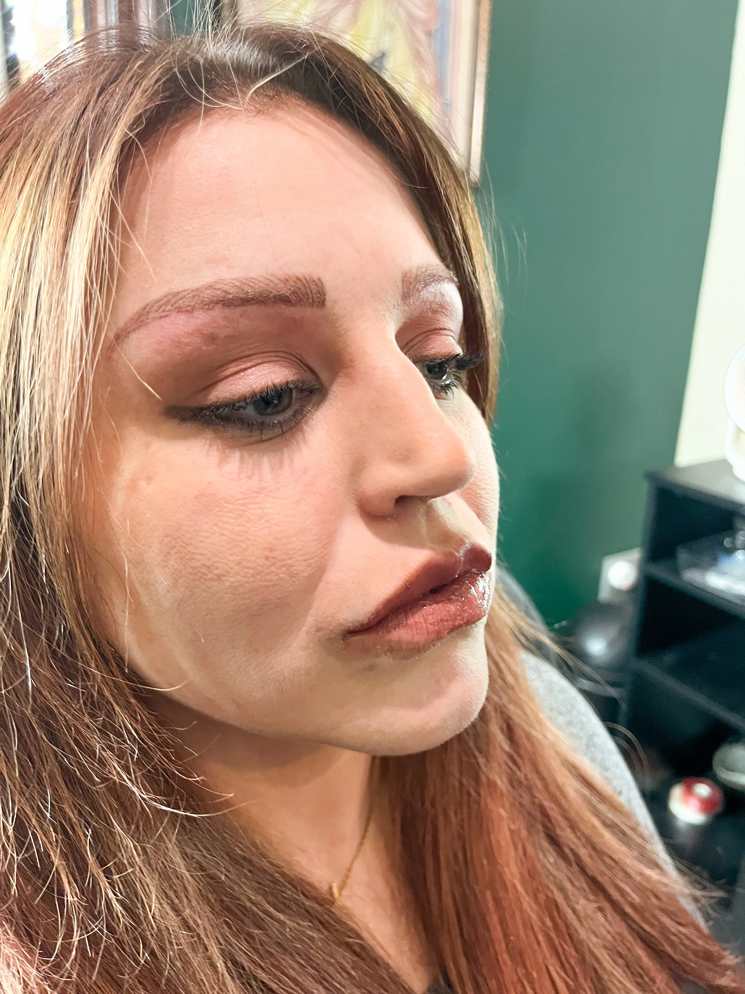 Close-up of a woman with long reddish-brown hair, makeup, and a neutral expression, in a salon or similar setting.