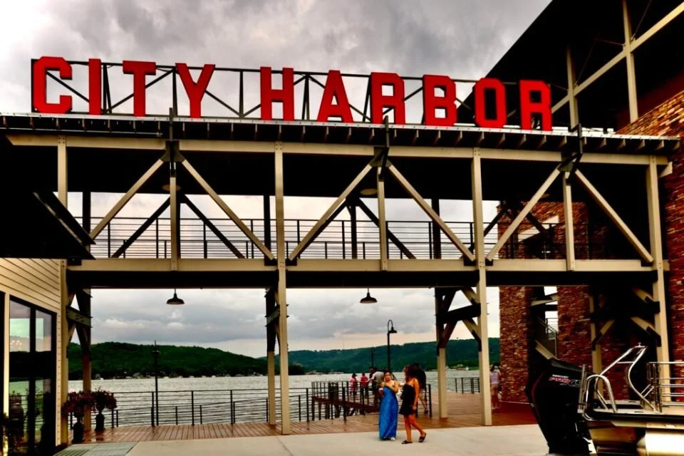 The sign reads 'City Harbor' on a metal structure above a waterfront promenade on Lake Guntersville with people walking, overlooking a harbor with hills in the background.