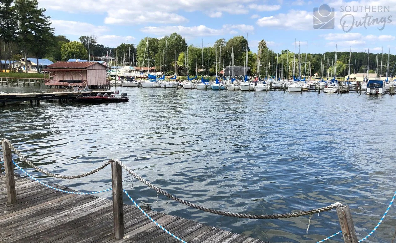 Guntersville marina with numerous sailboats docked, a wooden pier with a rope fence, and trees in the background on a partly cloudy day.