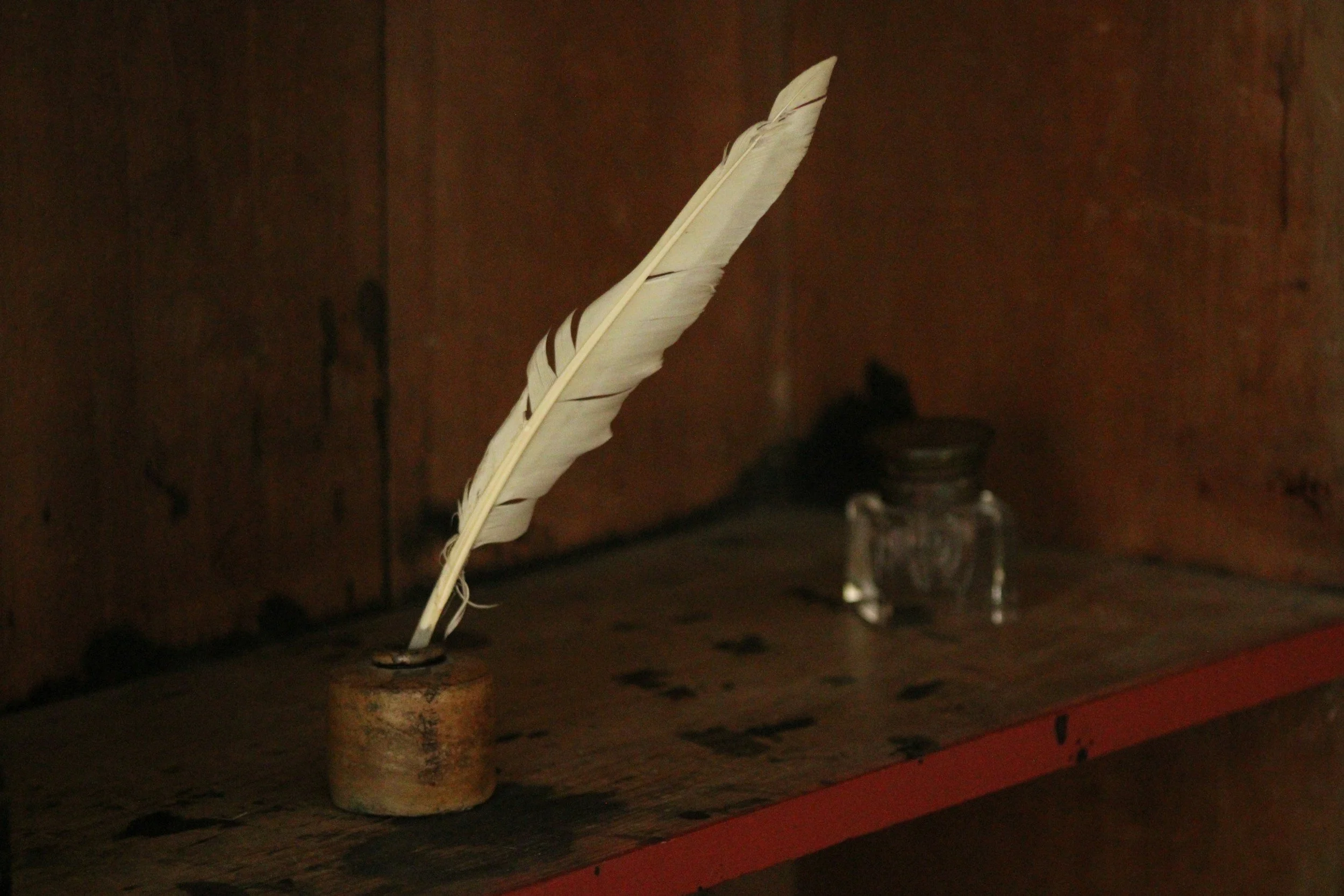 An off-white feather standing upright in a small container on a wooden surface, with a dark background and some glass objects in the distance.