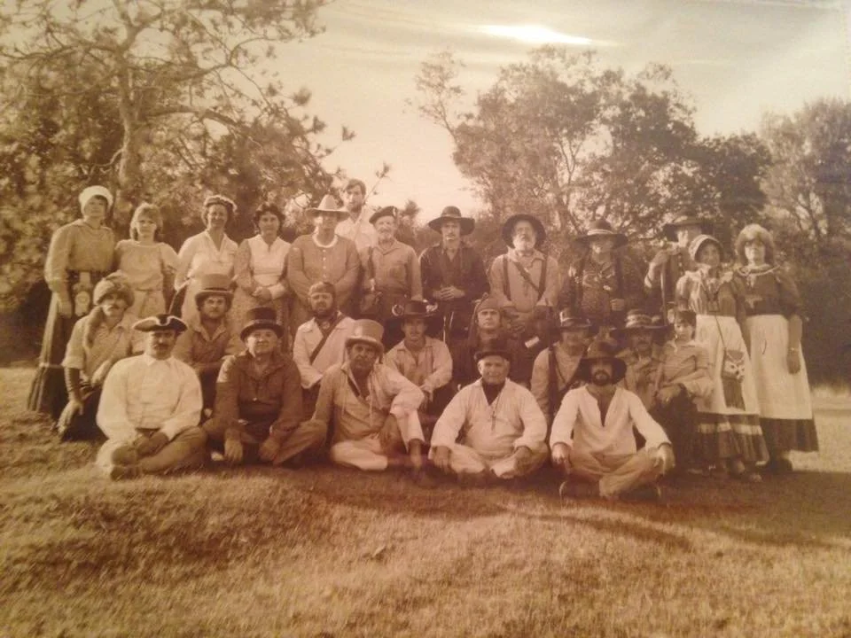 A sepia-toned vintage photo of a large group of men and women dressed in old-fashioned clothing, posing outdoors on grass with trees in the background.