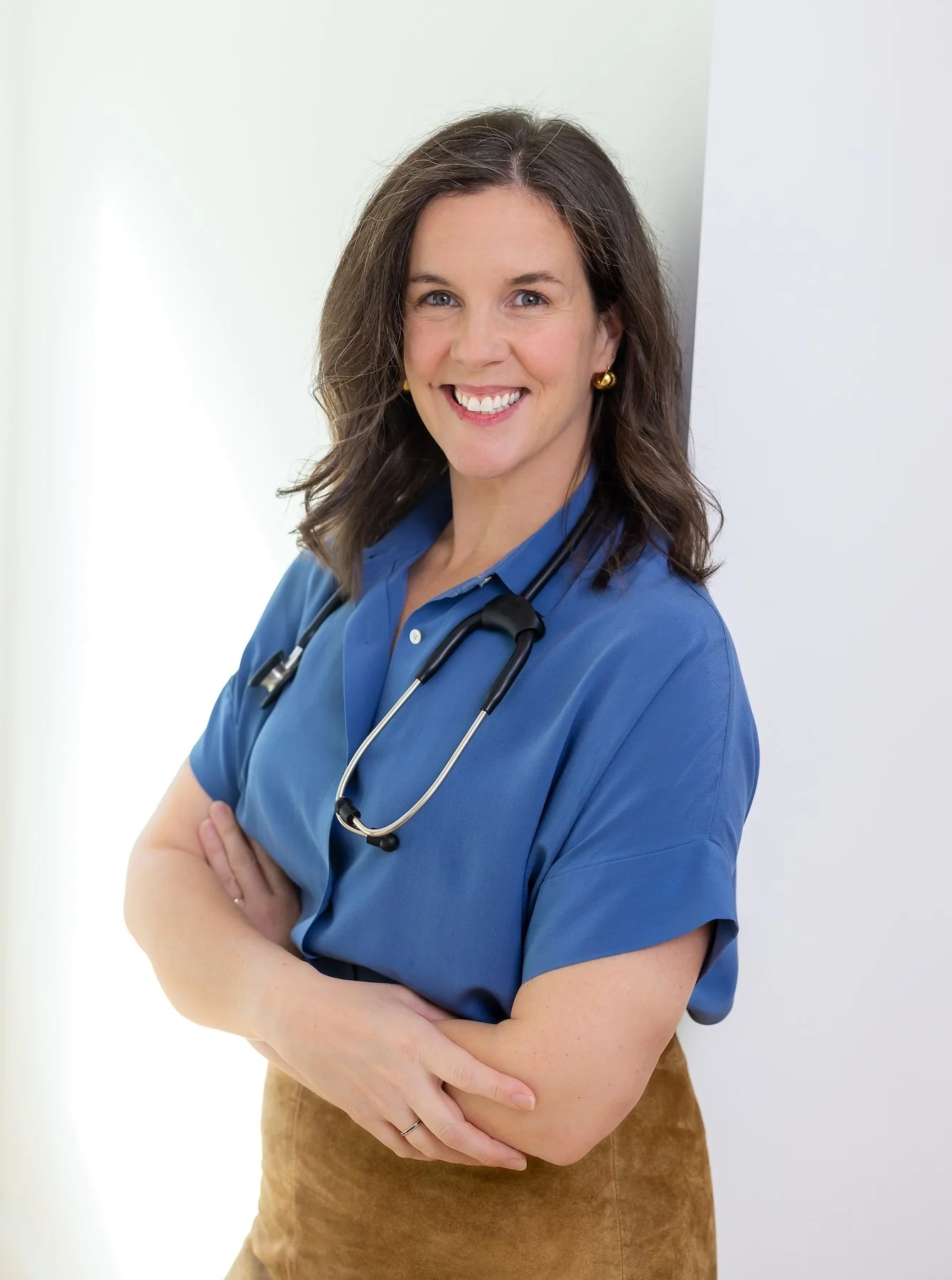 A female healthcare professional with shoulder-length brown hair, smiling, wearing blue scrubs and a stethoscope around her neck, standing with arms crossed against a neutral background.