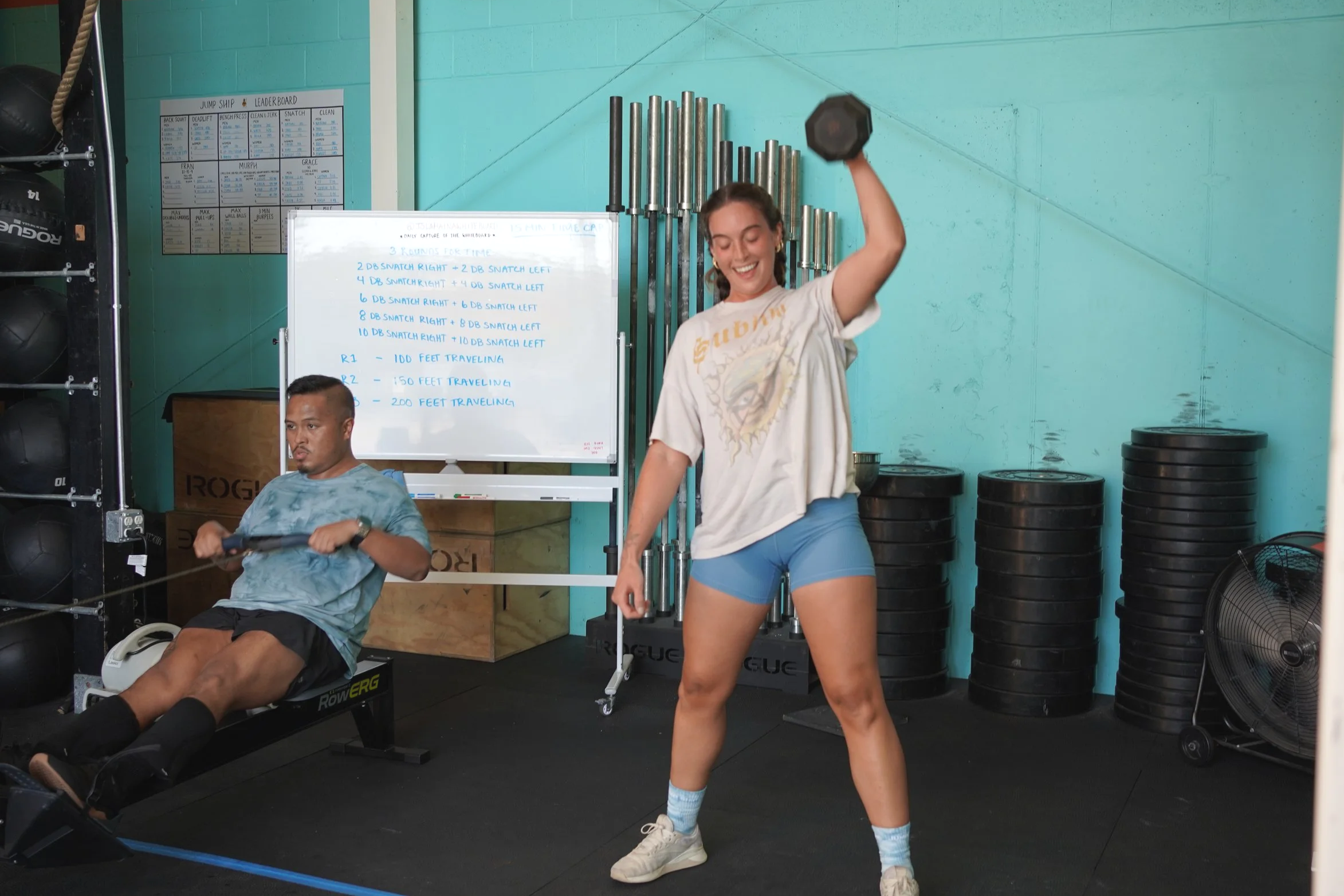 A woman lifting a dumbbell in a gym, smiling, while a man uses a rowing machine nearby. There are weights stacked against the wall and a whiteboard with workout instructions in the background.