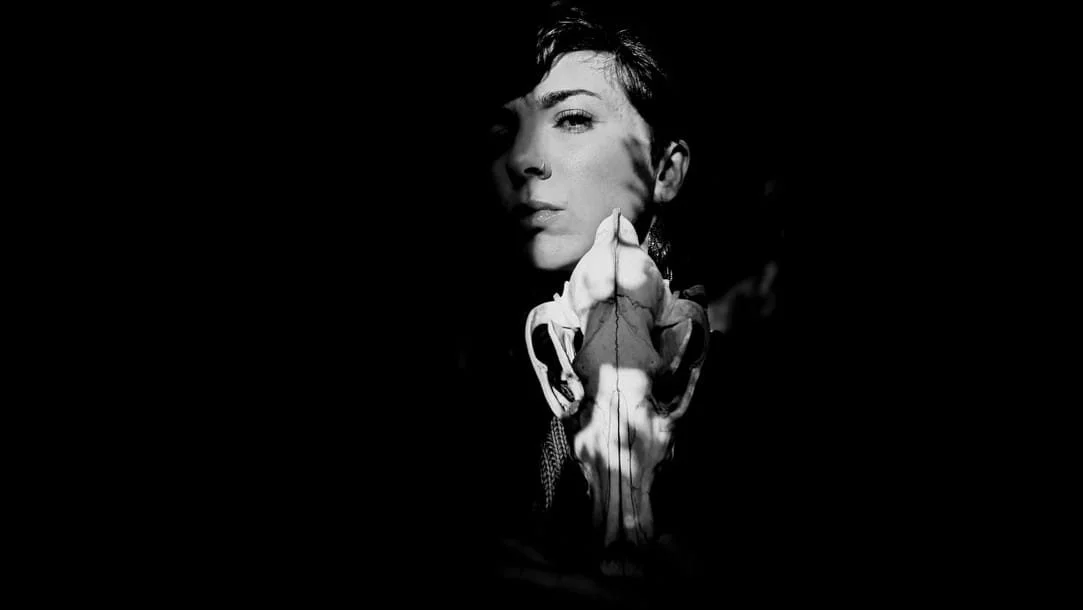 A black and white photo of a woman's face with short hair, holding a skull with her hand, creating a shadow that resembles a skull on her face.