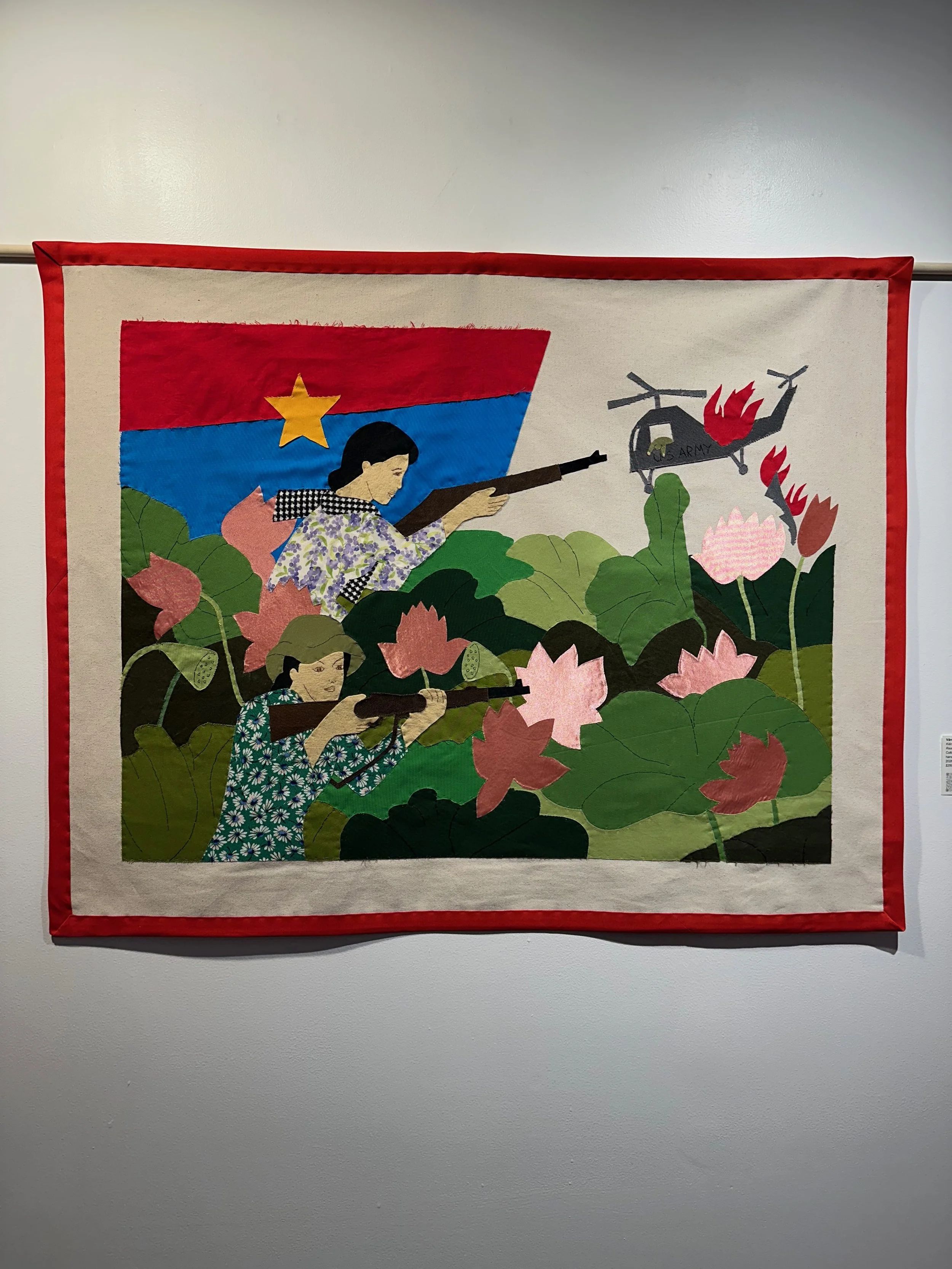 Folk art quilt depicting women with guns in a lotus pond, with a flag in the background, surrounded by a red border.
