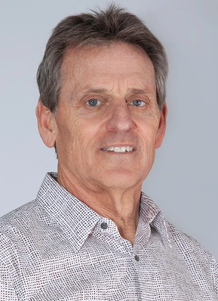 A middle-aged man with gray hair and blue eyes, wearing a white button-up shirt with small black dots, standing against a light gray background.