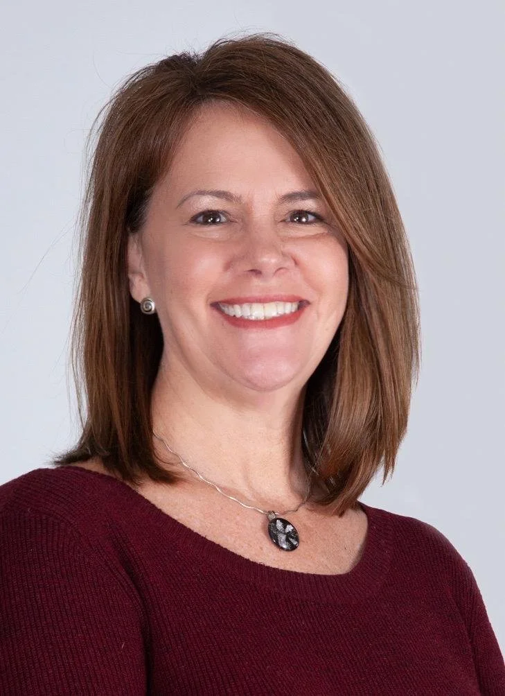 A woman with shoulder-length reddish-brown hair, smiling, wearing a burgundy top, earrings, and a necklace with a black and silver pendant.