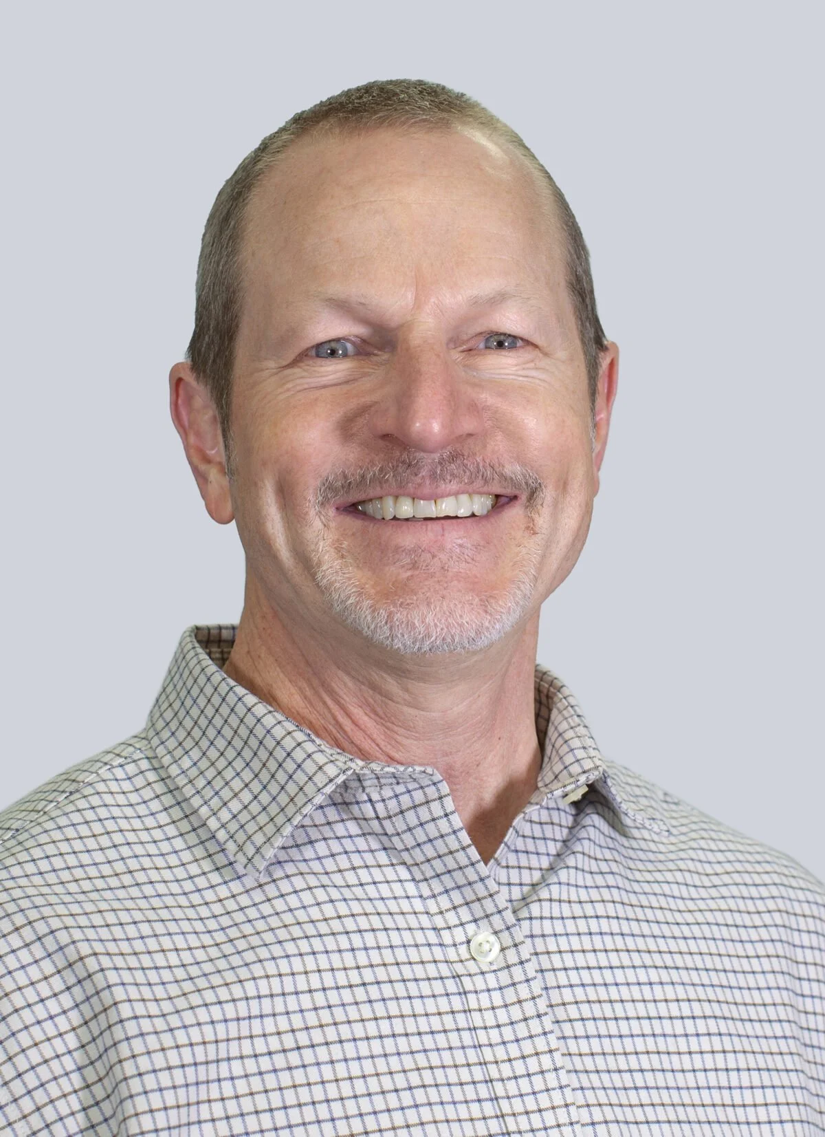 A middle-aged man with short, light brown hair and a goatee, wearing a checkered button-up shirt, smiling against a light gray background.