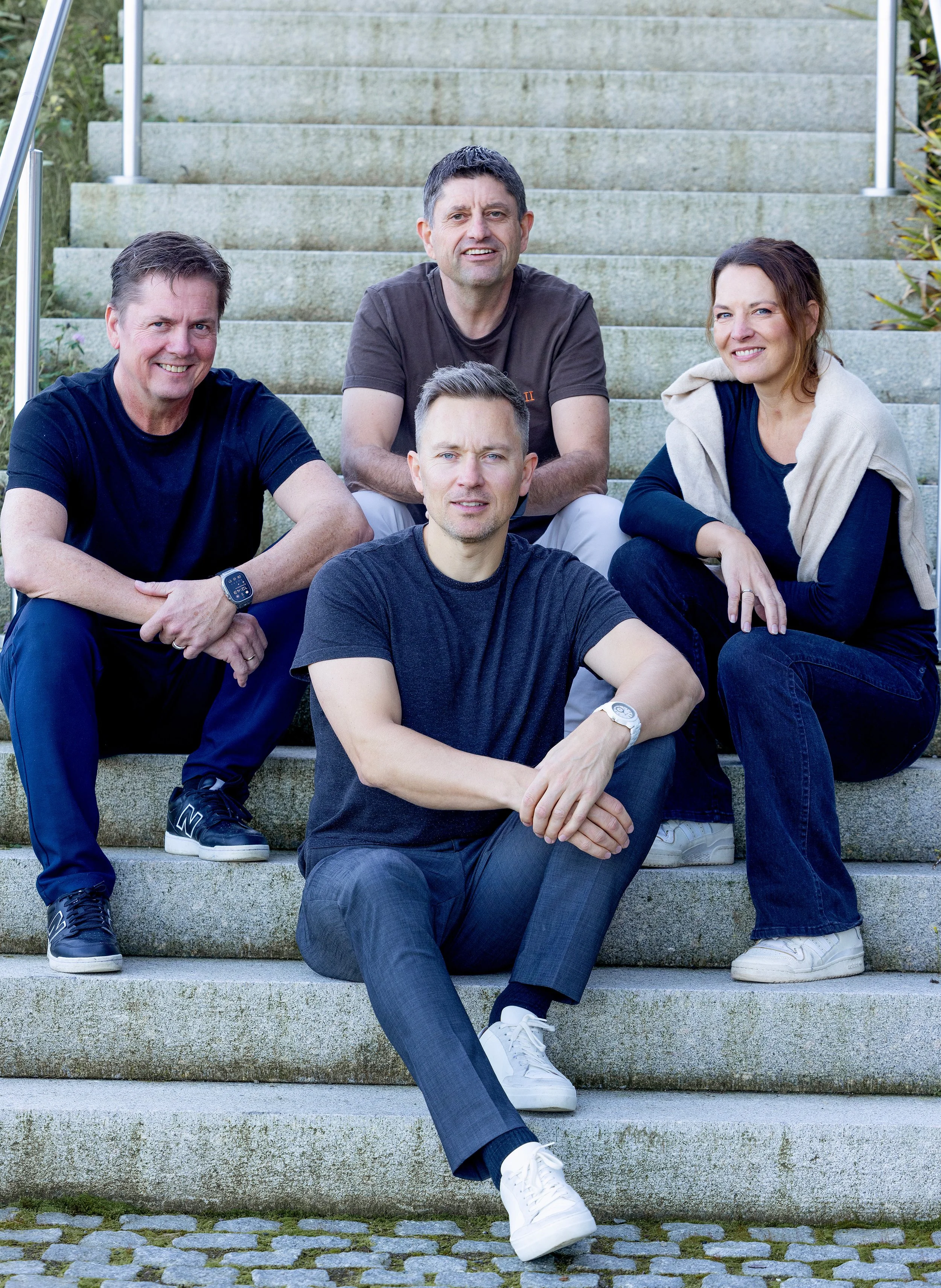 Fem personer sittende på trappetrinn utendørs, to menn og to kvinner, med smil og avslappet holdning