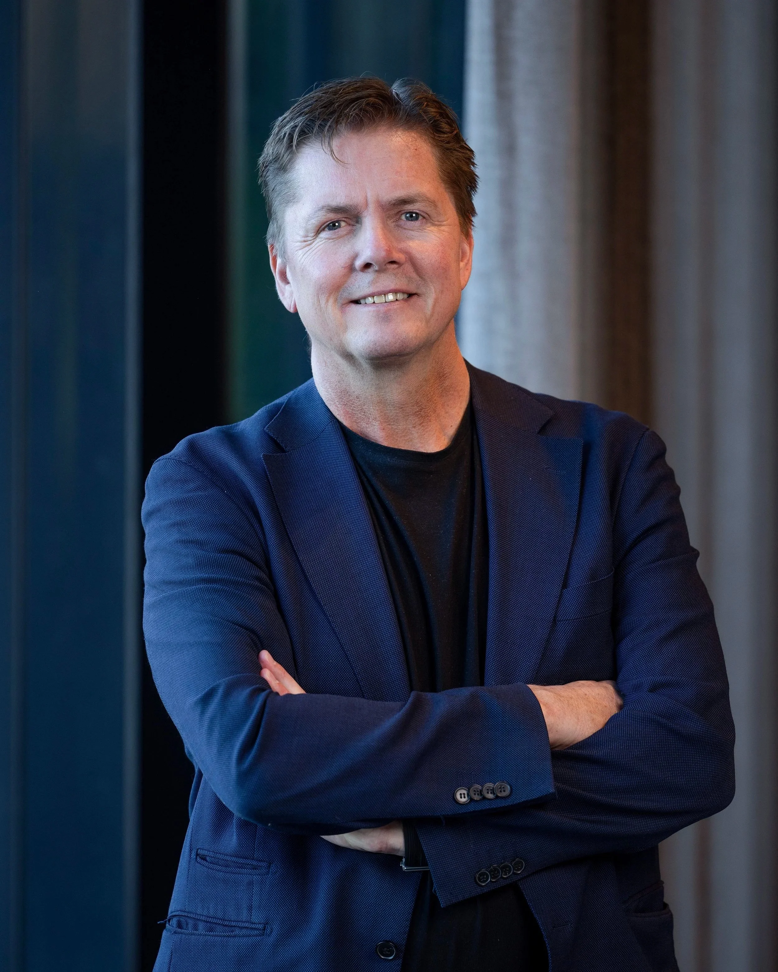 Mann med blå dress og svart skjorte står med armer crossed, smilende, i innendørs setting.