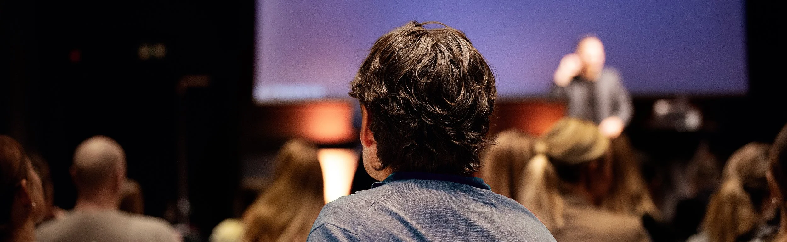 Person med brunt, krøllete hår som ser på en presentasjon eller foredrag med en stor skjerm foran seg på en konferanse eller seminar.