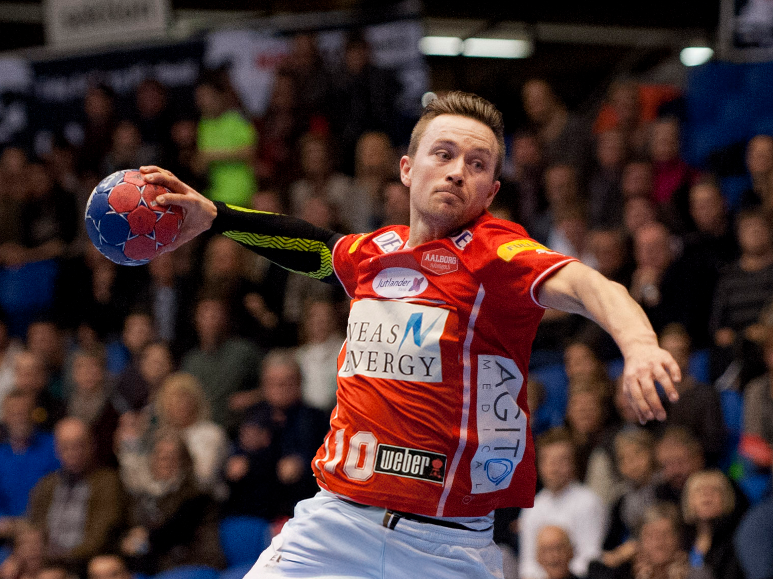 En mann som spiller håndball, kledd i oransje drakt og har på seg svart og grønn armvarmer, med publikum i bakgrunnen.