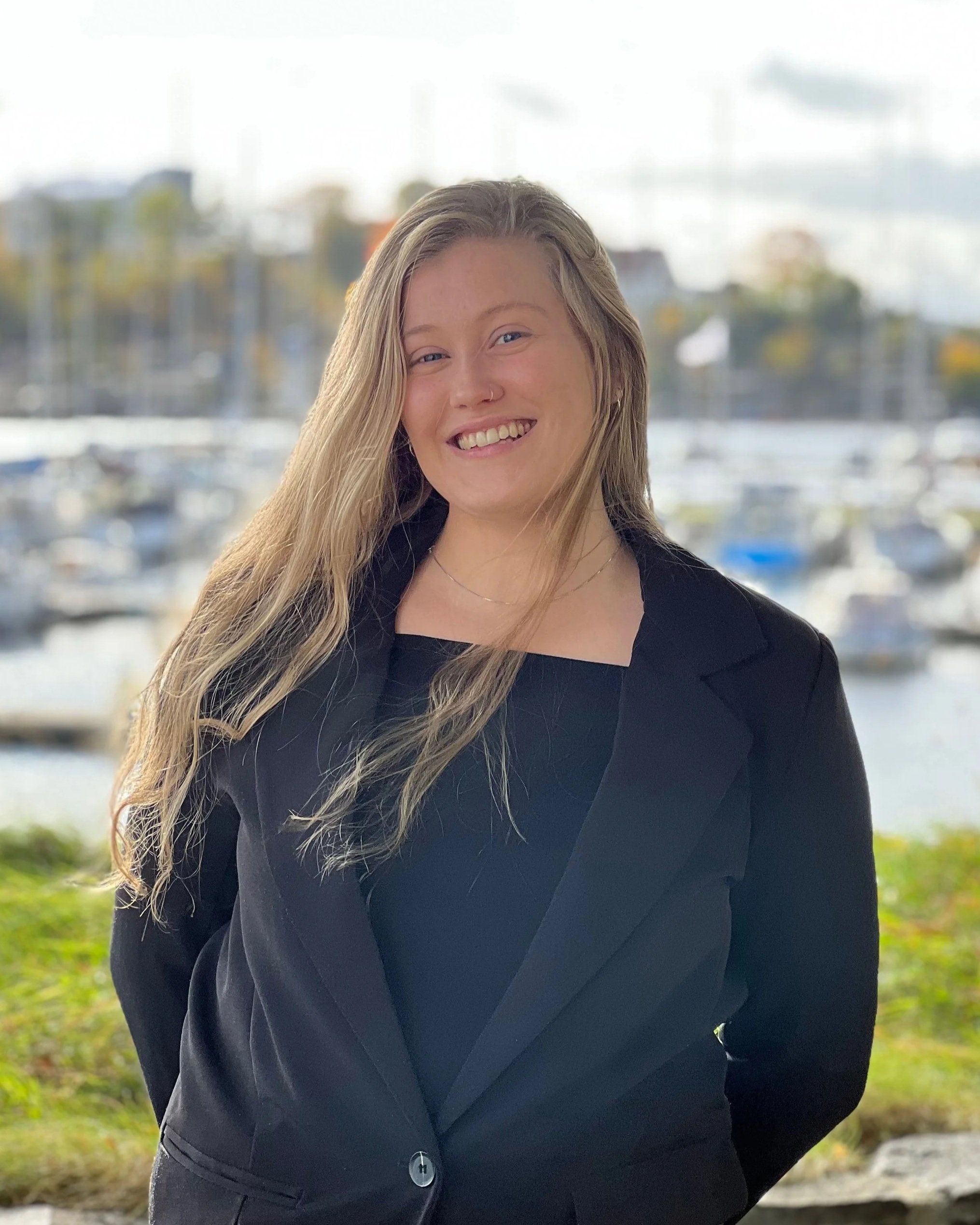 Smilende kvinne med langt, blond hår og svart klær, stående utendørs med lyst, uskarpt vann og båter i bakgrunnen.