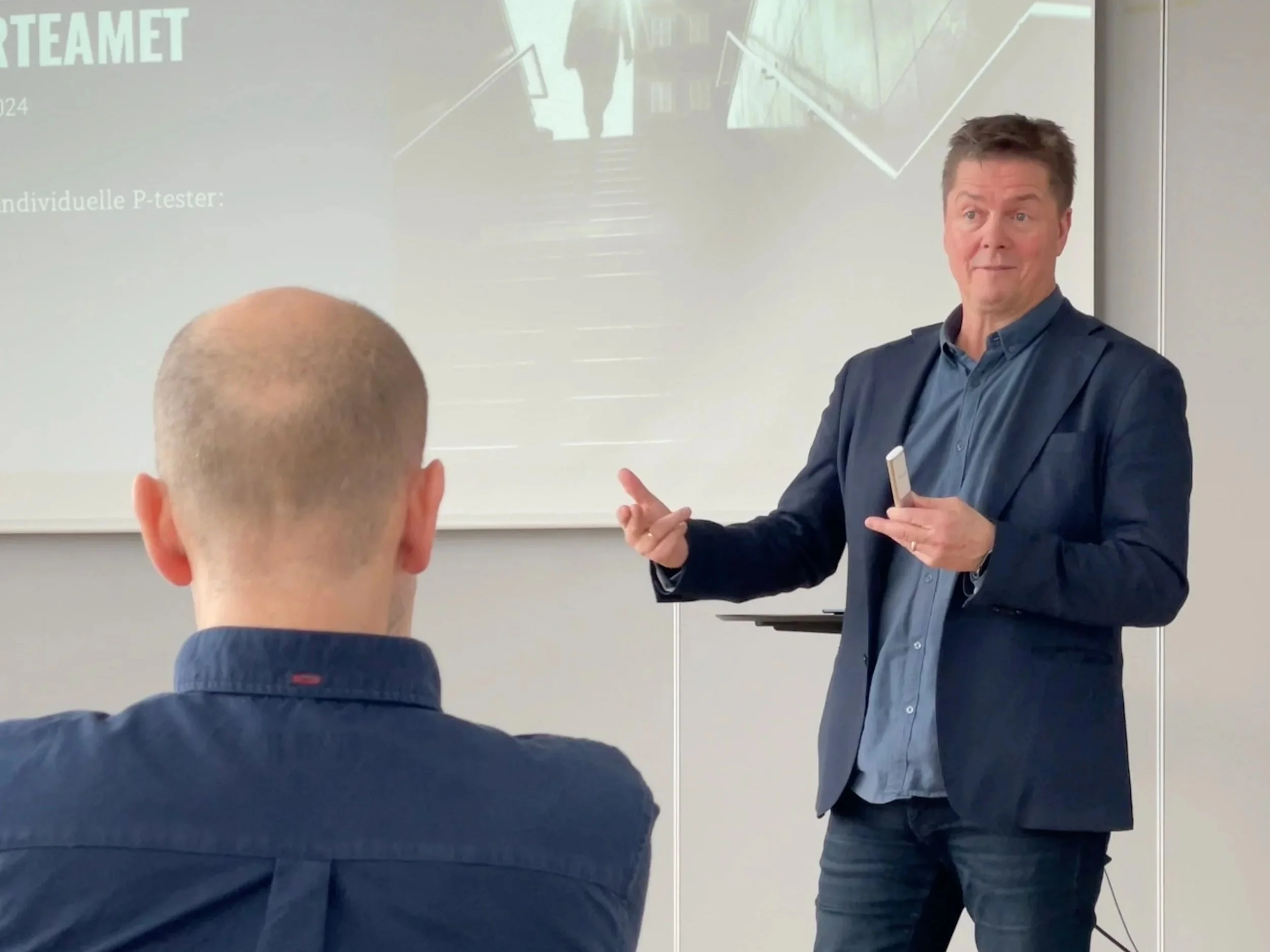 En mann som holder en tusjpenn og snakker med en annen person i en klasserom eller møterom, foran en skjerm med presentasjonstekst