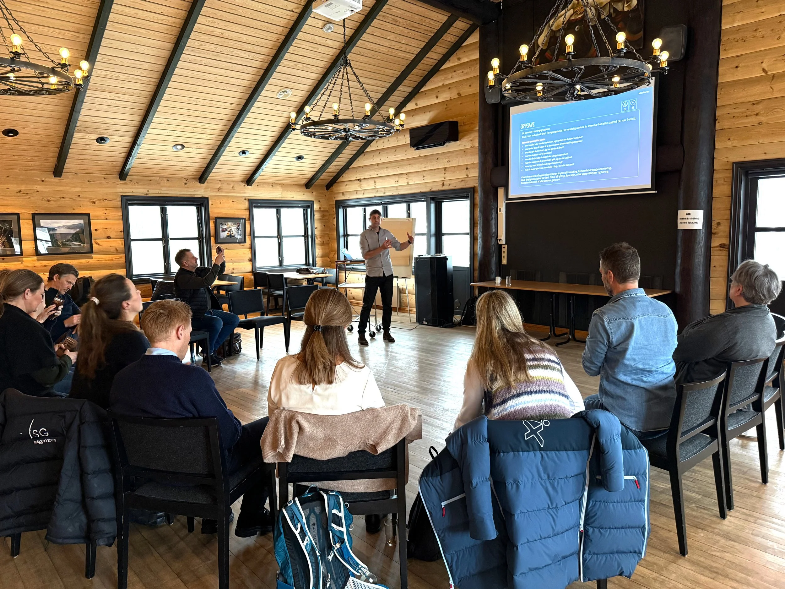 En gruppe mennesker deltar i en presentasjon i et rom med trebjelker og vegger. En man står foran en skjerm med presentasjonsinformasjon, mens andre sitter i stoler og noen tar bilder.