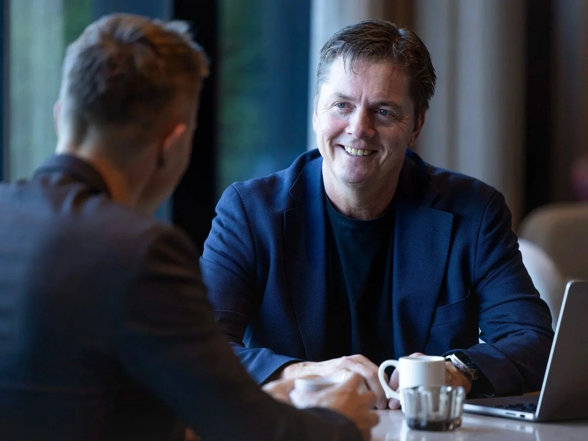 To menn sitter ved et bord og har samtale, med en kopp og en laptop på bordet.