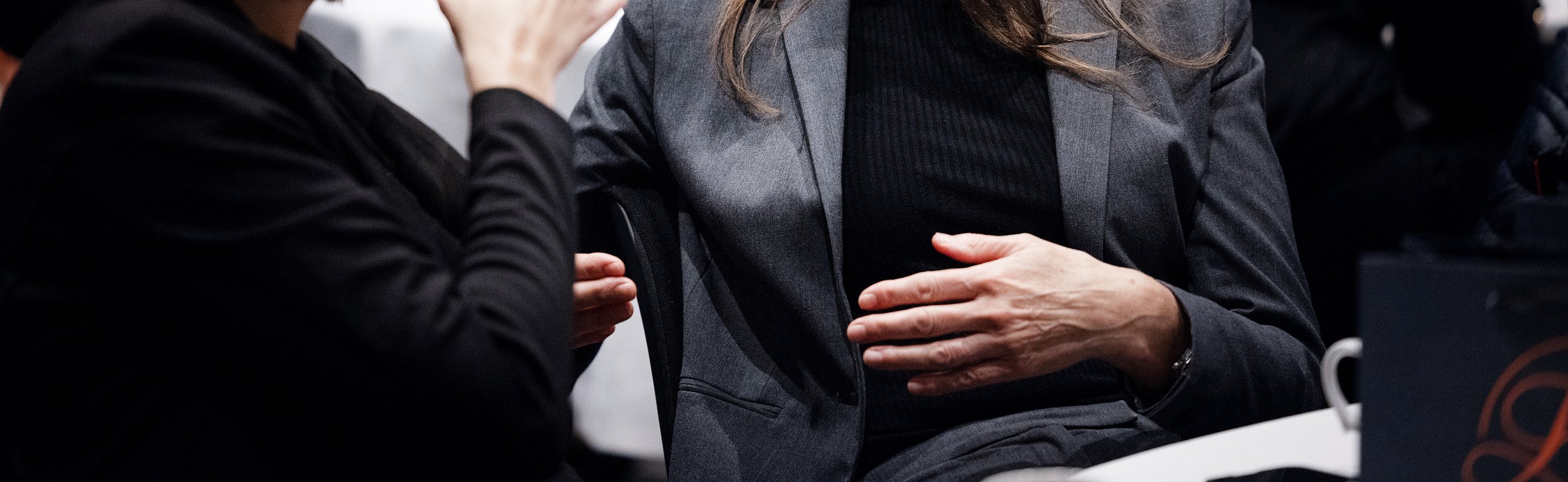 To personer som snakker, en med svart klær og en med grå jakke, innendørs.