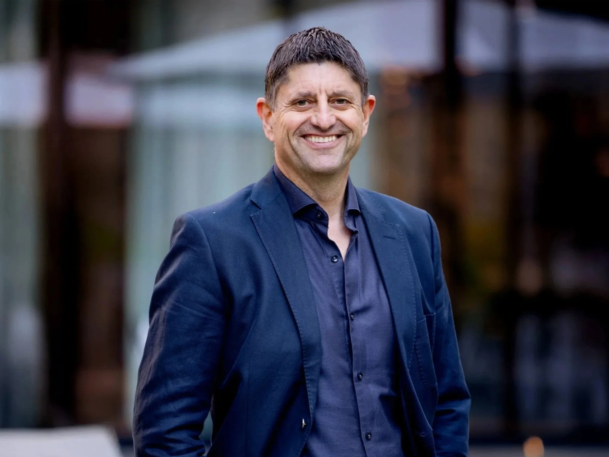 En fornøyd mann med kort brunt hår, iført mørkeblå dress, står utendørs med en uskarp bakgrunn av tre og bygg.