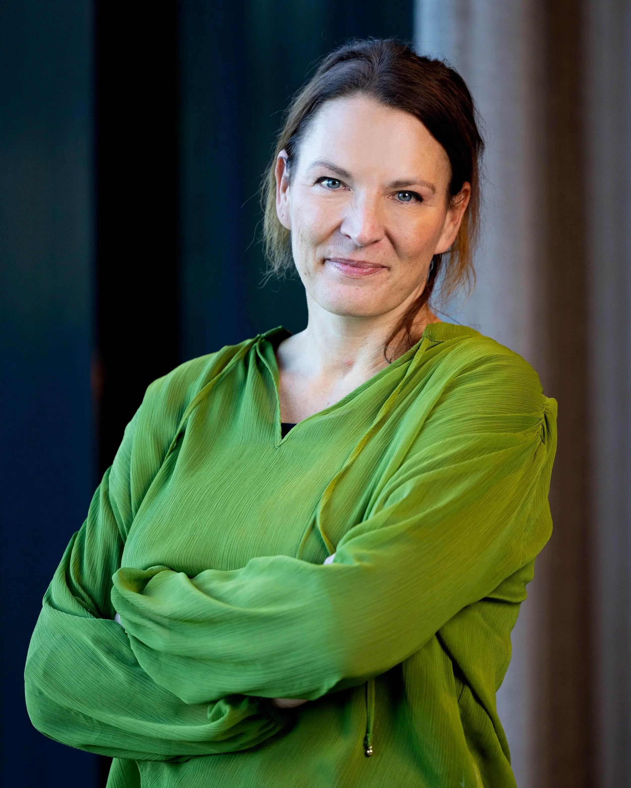 Portrett av en kvinne med kort, mørk hår og blå øyne, som har på seg en grønn, langermet kjole, med armene krysset, står foran en mørk og lys bakgrunn.