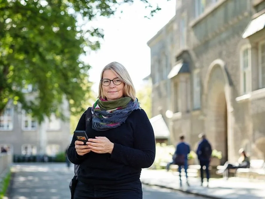 En kvinne står på gaten med en smarttelefon, omgitt av trær og eldre bygninger, og smiler.