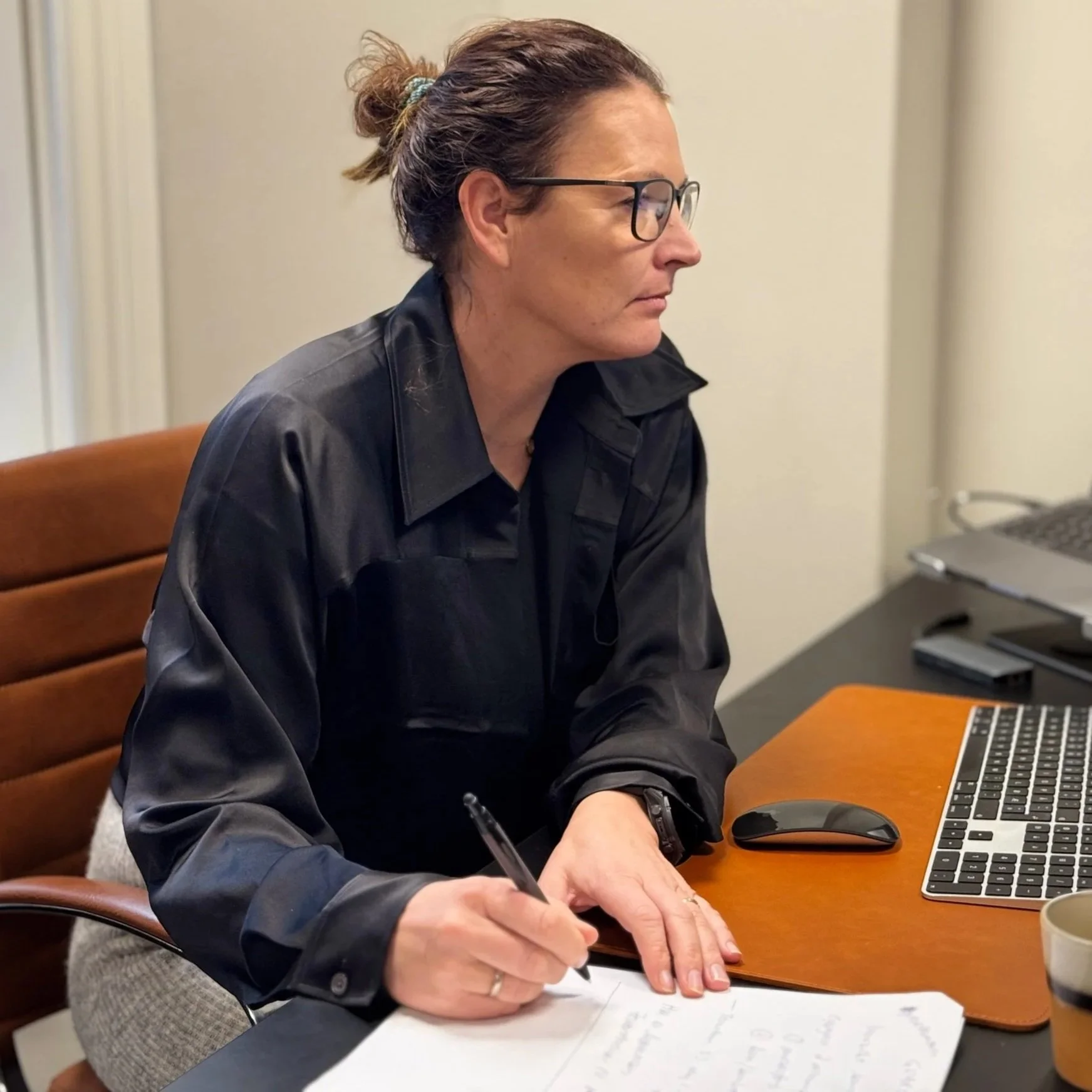 Kvinne med briller skriver på papir ved skrivebord med datamaskin i et kontor.