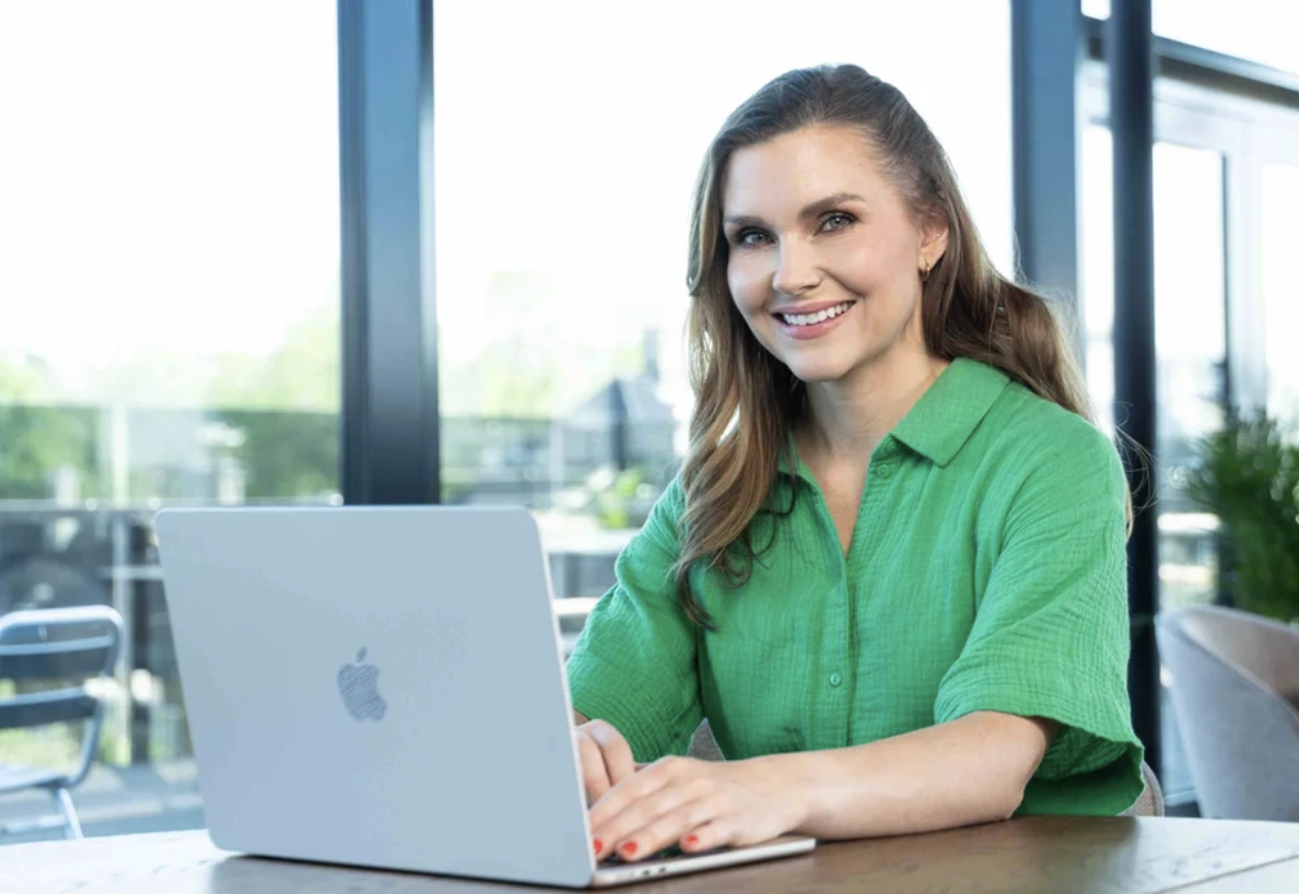 En smilende kvinne med langt brunt hår, iført en grønn skjorte, som sitter ved et bord med en åpen MacBook i en moderne kontorbygning med store vinduer.