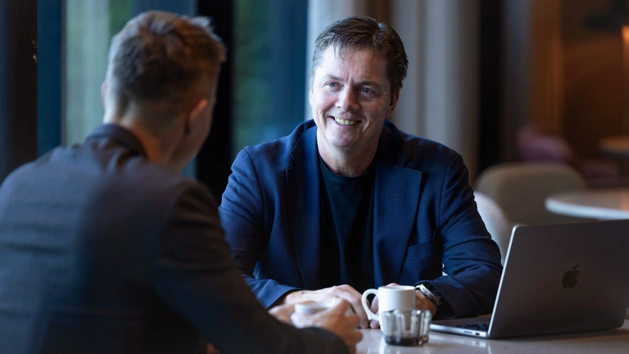 To menn sitter ved et kaffebord og snakker i et rom med store vinduer, det er en hyggelig samtale, og en av dem smiler.
