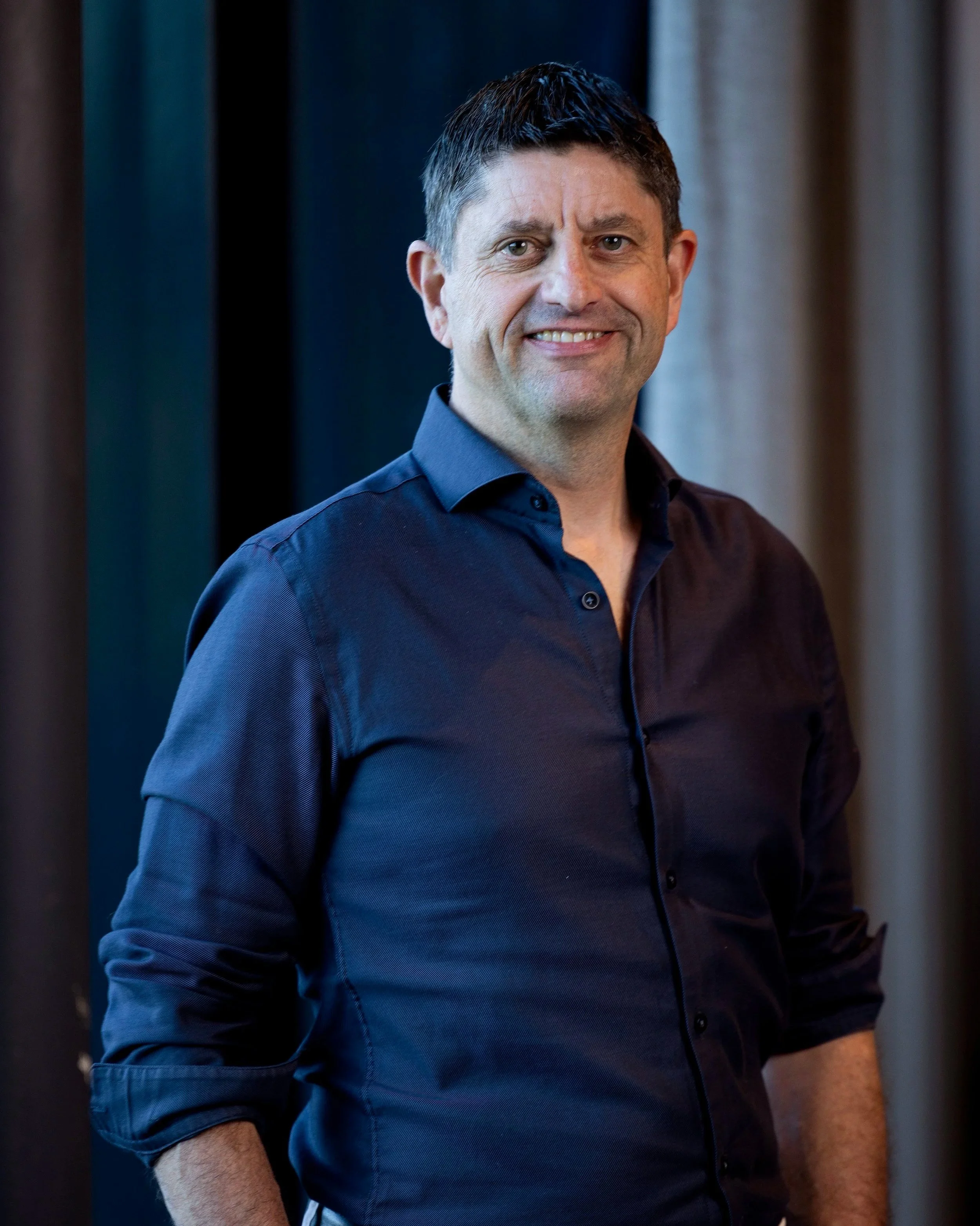 Portrett av en smilende mann med kort mørkt hår, iført en mørk blå skjorte, mot en bakgrunn med gardiner i forskjellige nyanser.