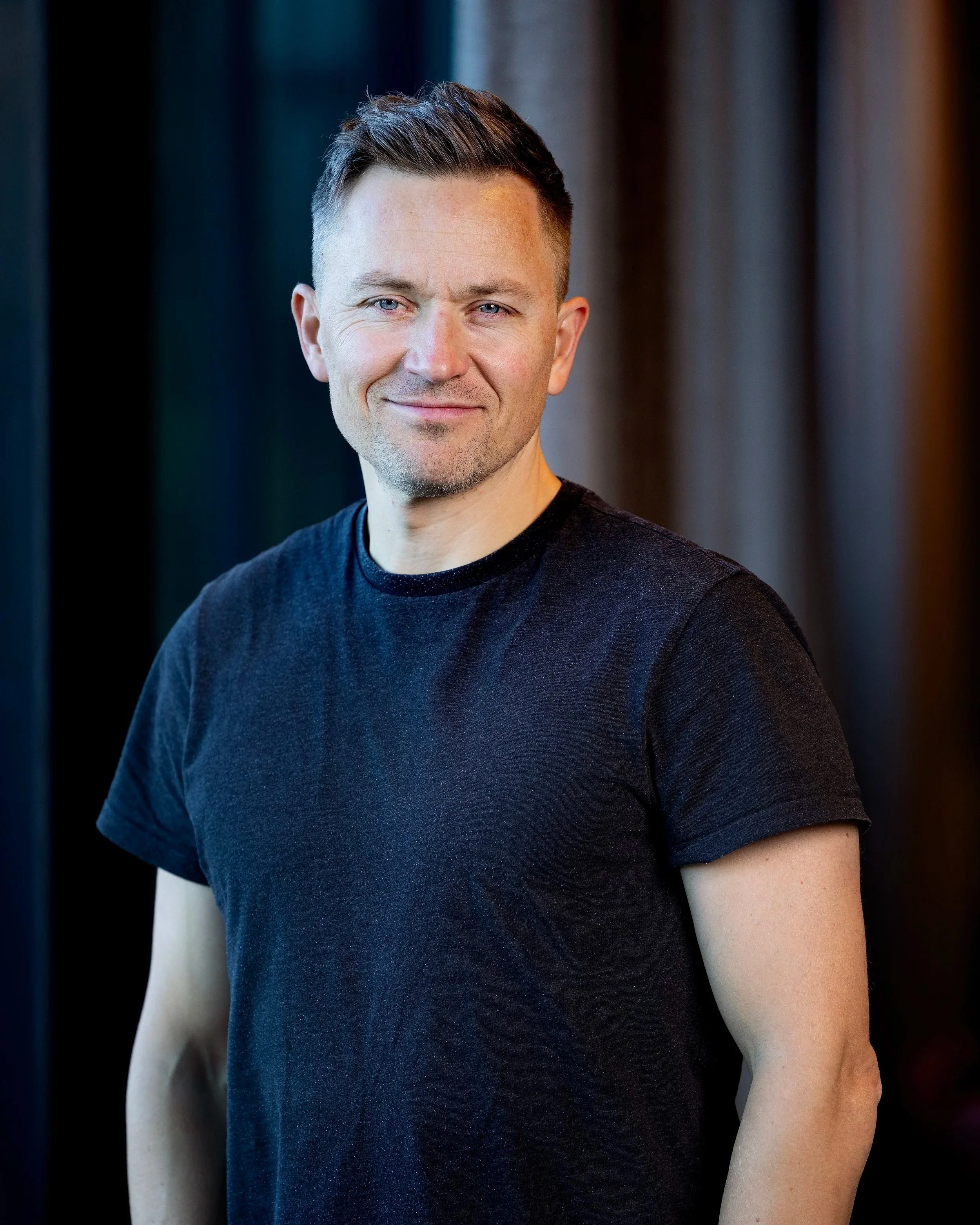 Nærbilde av en mann med kort, mørkt hår og blek hud, som smiler svakt og bærer en svart t-skjorte. Bakgrunnen er uklar, med mørke og nøytrale farger.