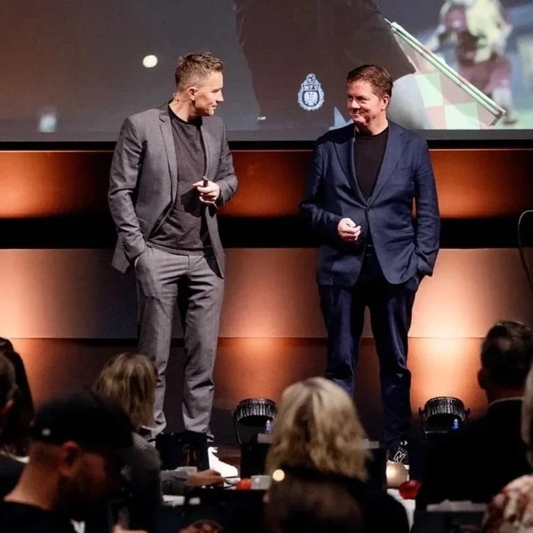 To menn i dresser snakker på en scene foran et publikum i et konferanserom.