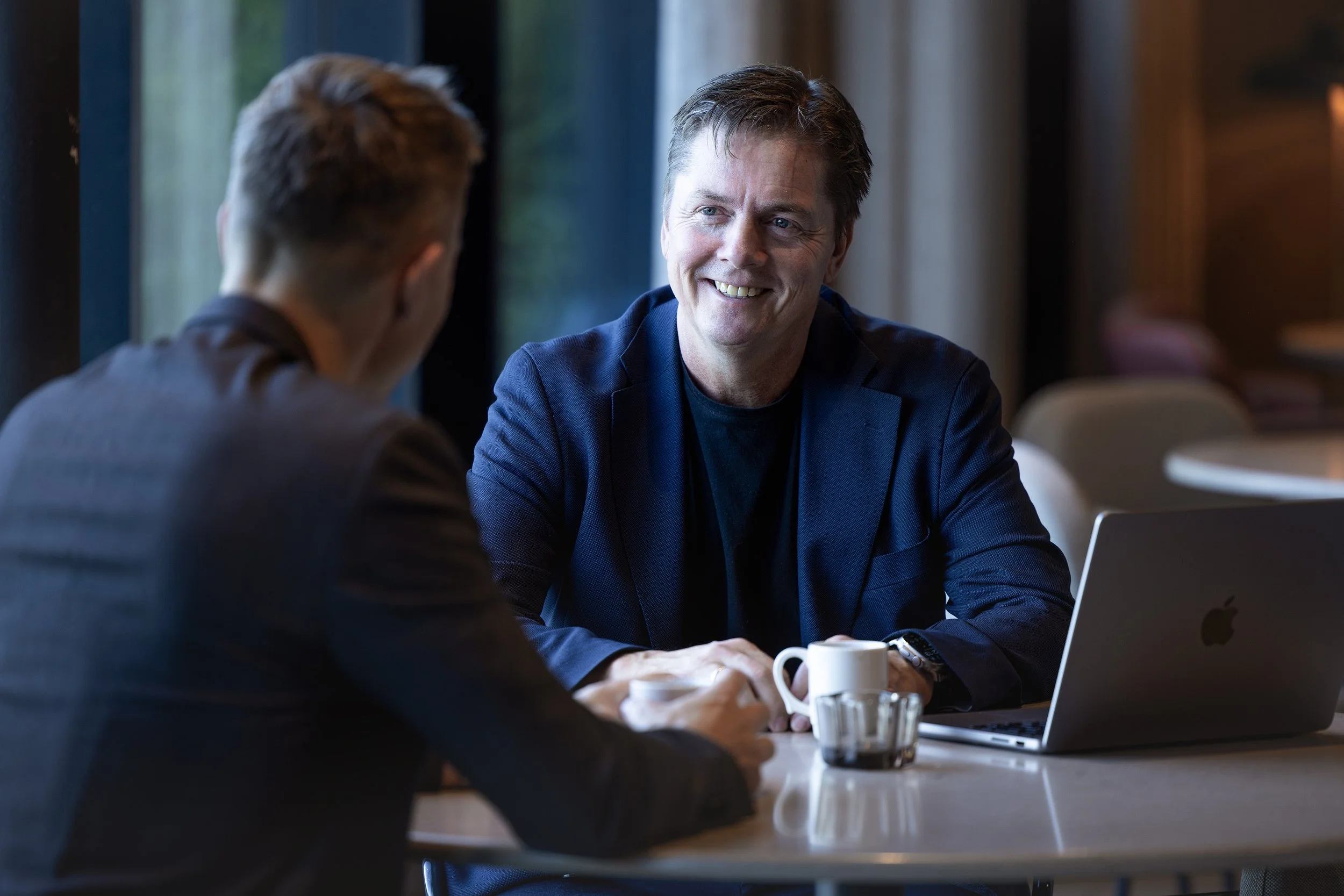 To menn snakker i et spiseområde, den ene er smilende og ser på den andre, med en åpen laptop foran dem, og kaffe kopper på bordet.