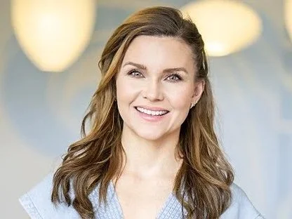 Et smilende kvinne med brunt, bølgete hår og lyse klær, bakgrunn med gule og hvite lys.