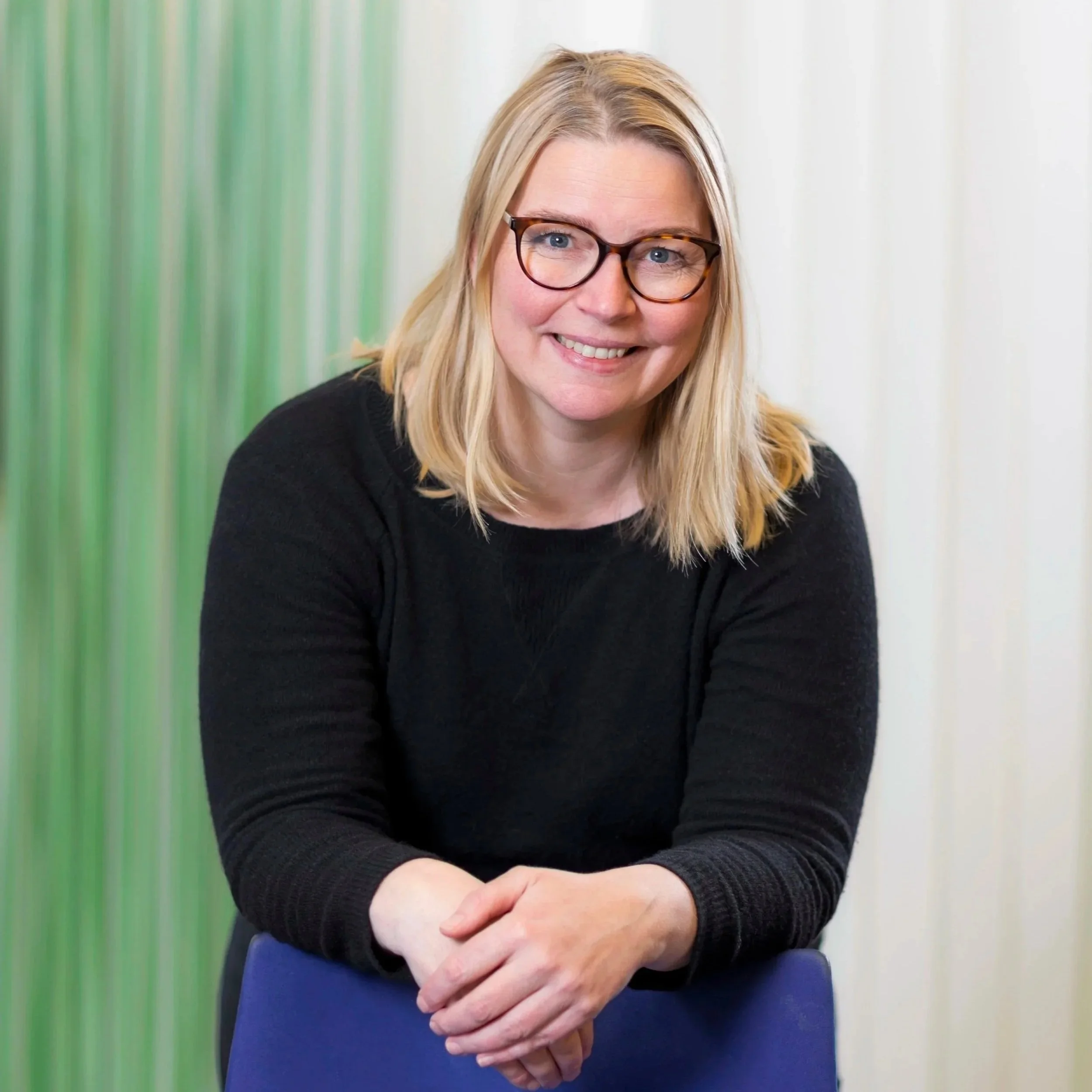 Nærbilde av en lykkelig kvinne med blondt, rett hår og briller, iført svart genser, som smilende ser mot kameraet med armene korslagt, foran en vegg med grønne og hvite striper.