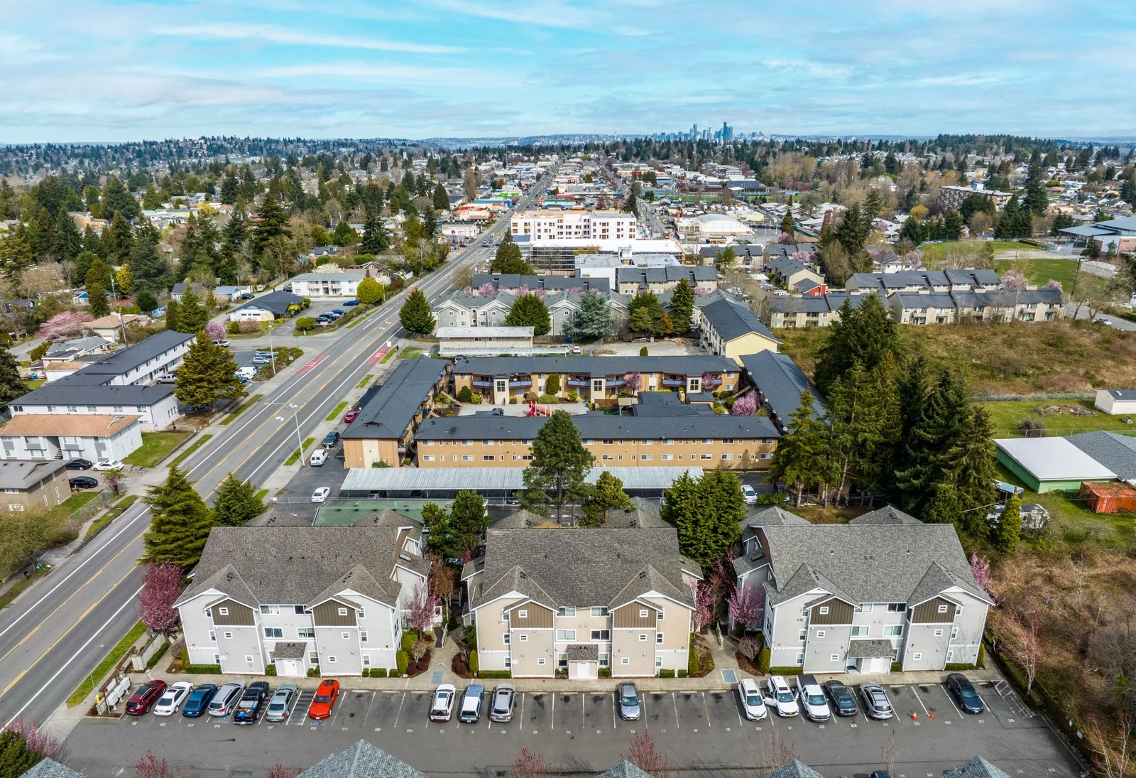 Aerial view of a city neighborhood with residential buildings, trees, and streets, extending to a distant downtown skyline under a partly cloudy sky.