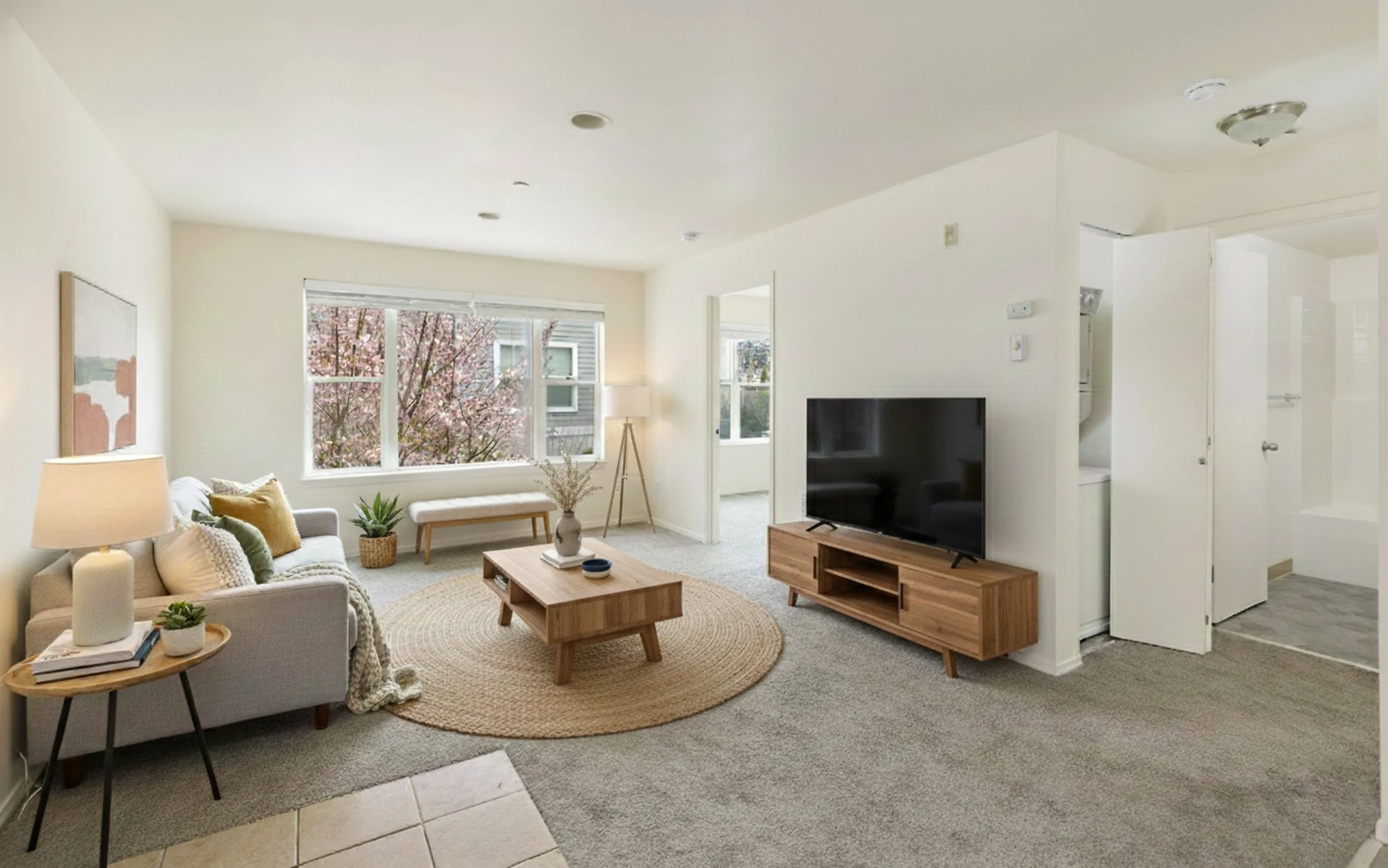 Bright living room with beige sofa, curly rug, wooden coffee table, TV on wooden stand, and large window with trees outside.