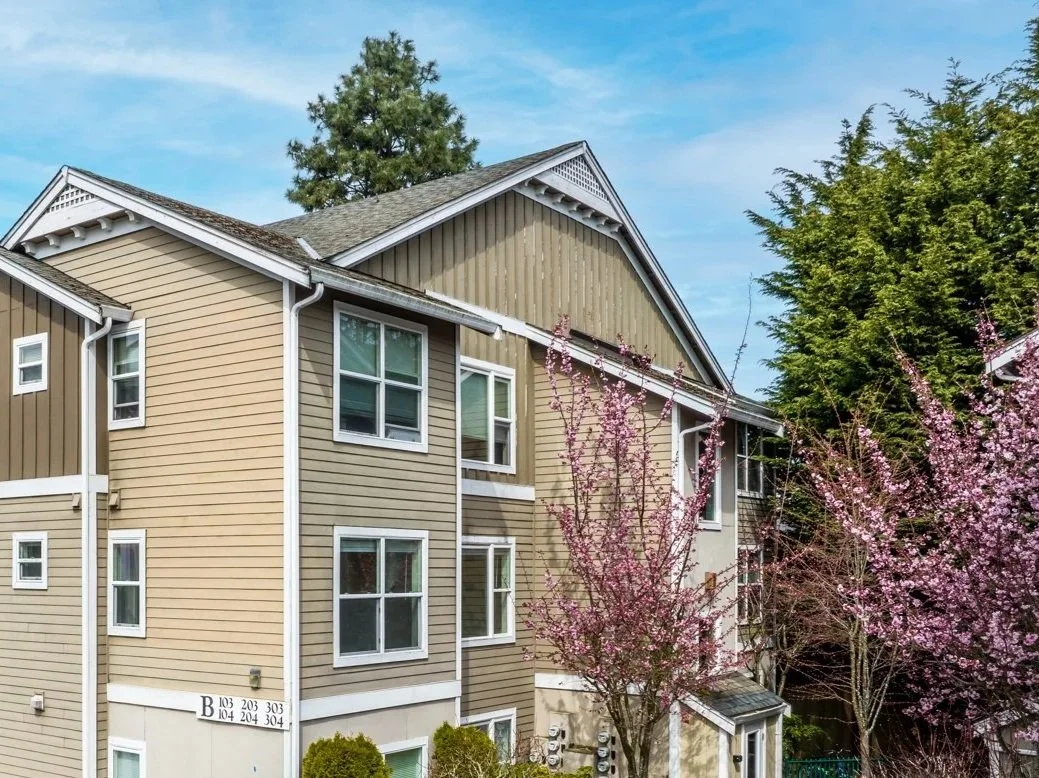 Multi-story residential building with beige and light brown siding, white window trim, surrounded by blooming pink trees and green foliage, under a blue sky.