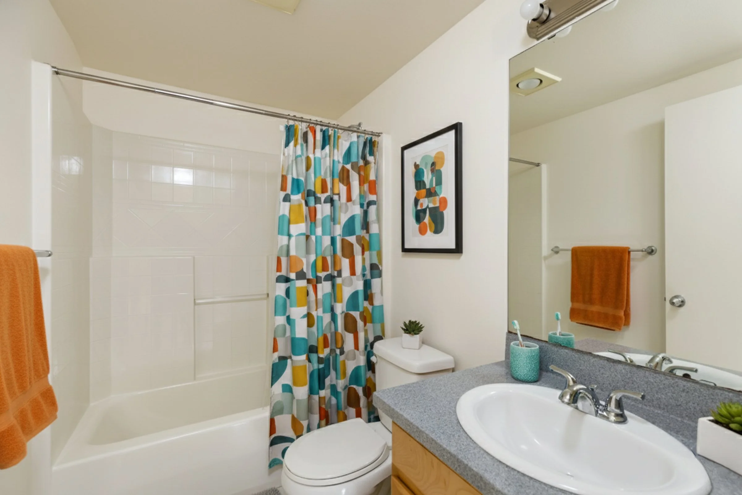 A bathroom featuring a white bathtub with a colorful patterned shower curtain, a white toilet with a small potted plant on top, a gray countertop with a white sink, toothbrush holders, a large mirror, and orange towels hanging on towel bars.