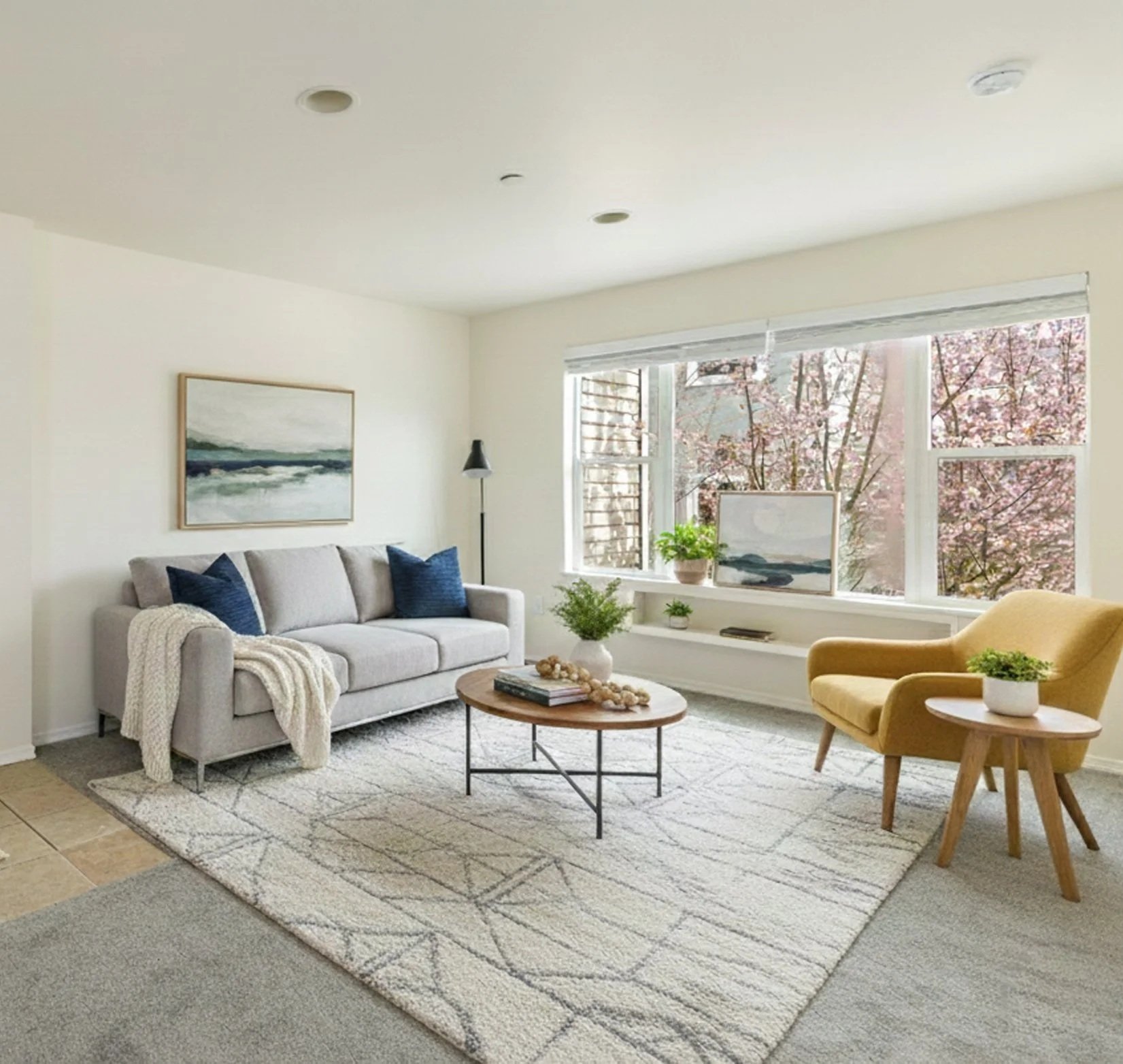A bright living room with a light gray sofa, blue accent pillows, a cream armchair, a round wooden coffee table, and a large window with cherry blossoms outside. Decor includes plants, artwork, and a neutral rug.