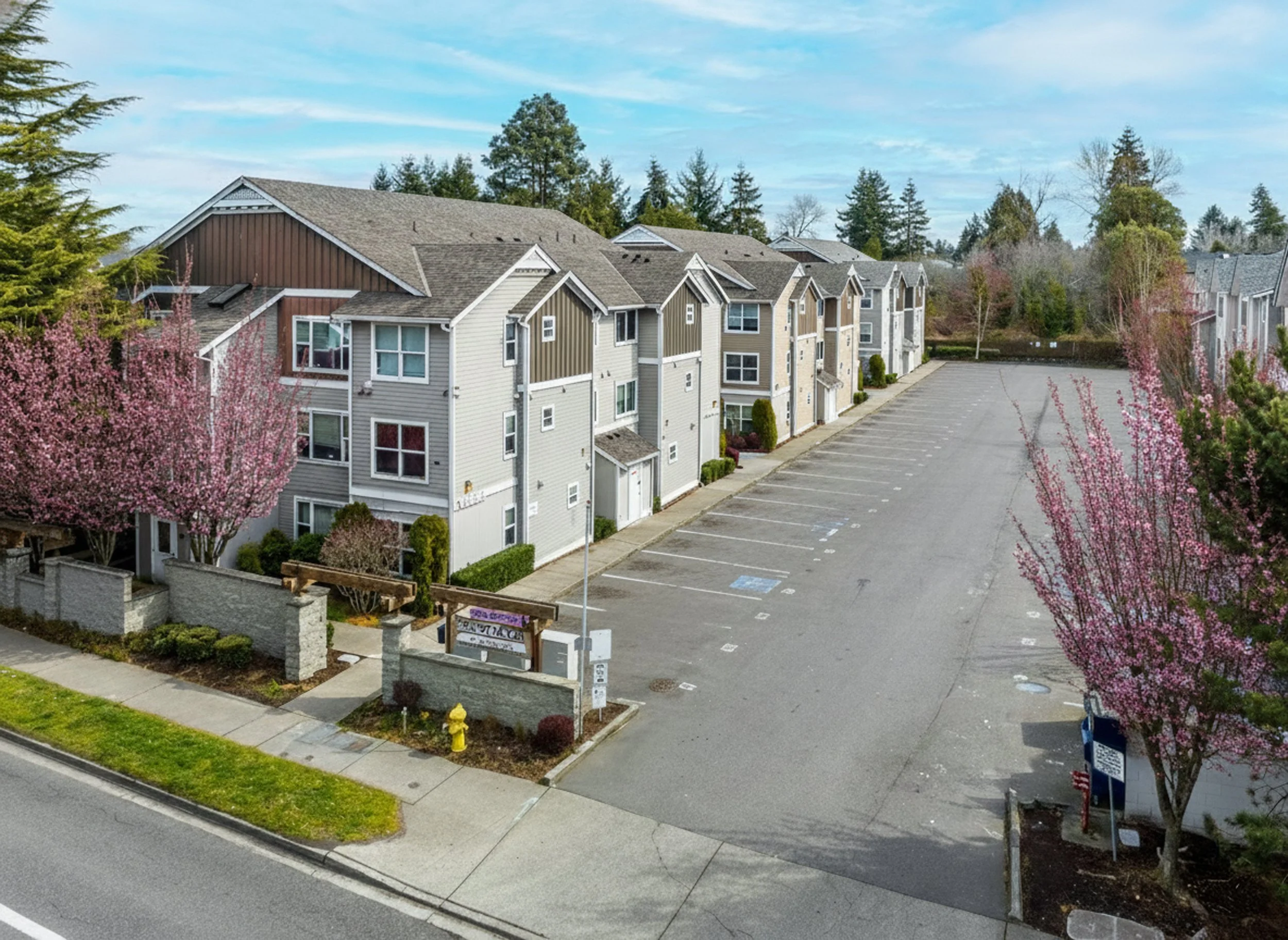 Apartment complex with pink blossom trees, empty parking lot, and clear sky.