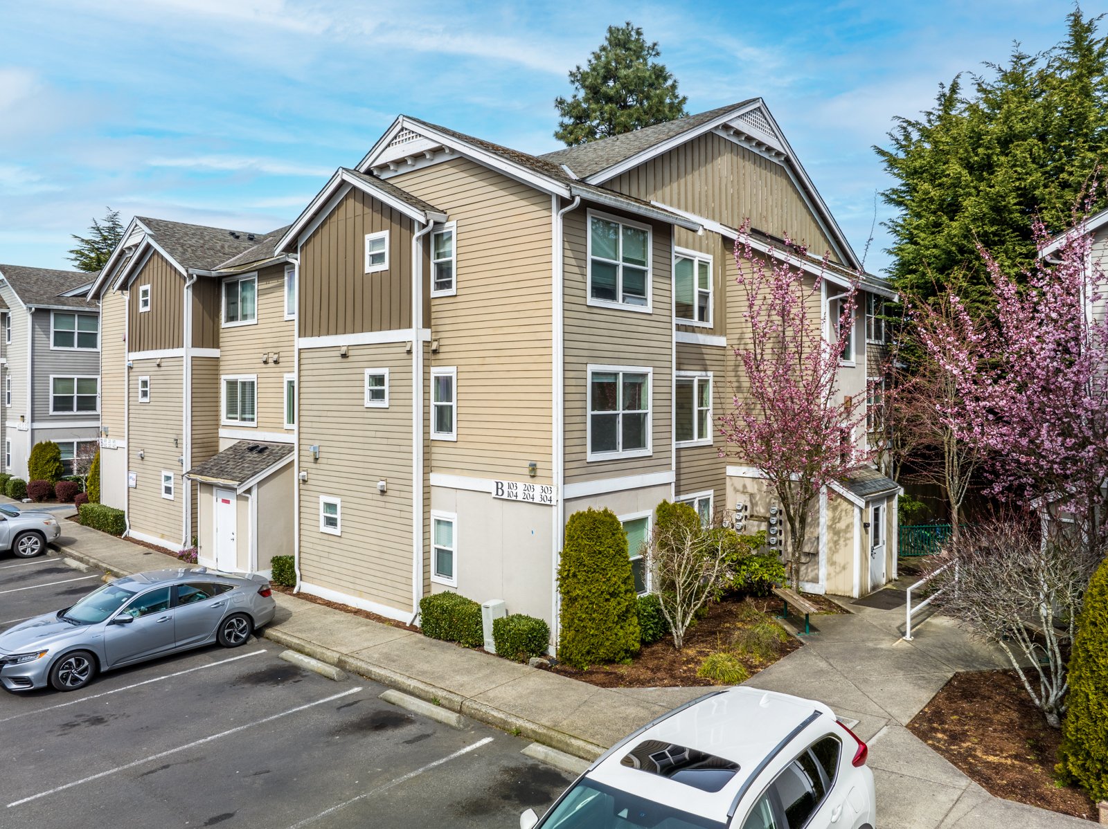 Multi-story residential apartment building with beige and brown exterior, white window frames, and pink flowering trees in front, with parking lot and cars near sidewalk.