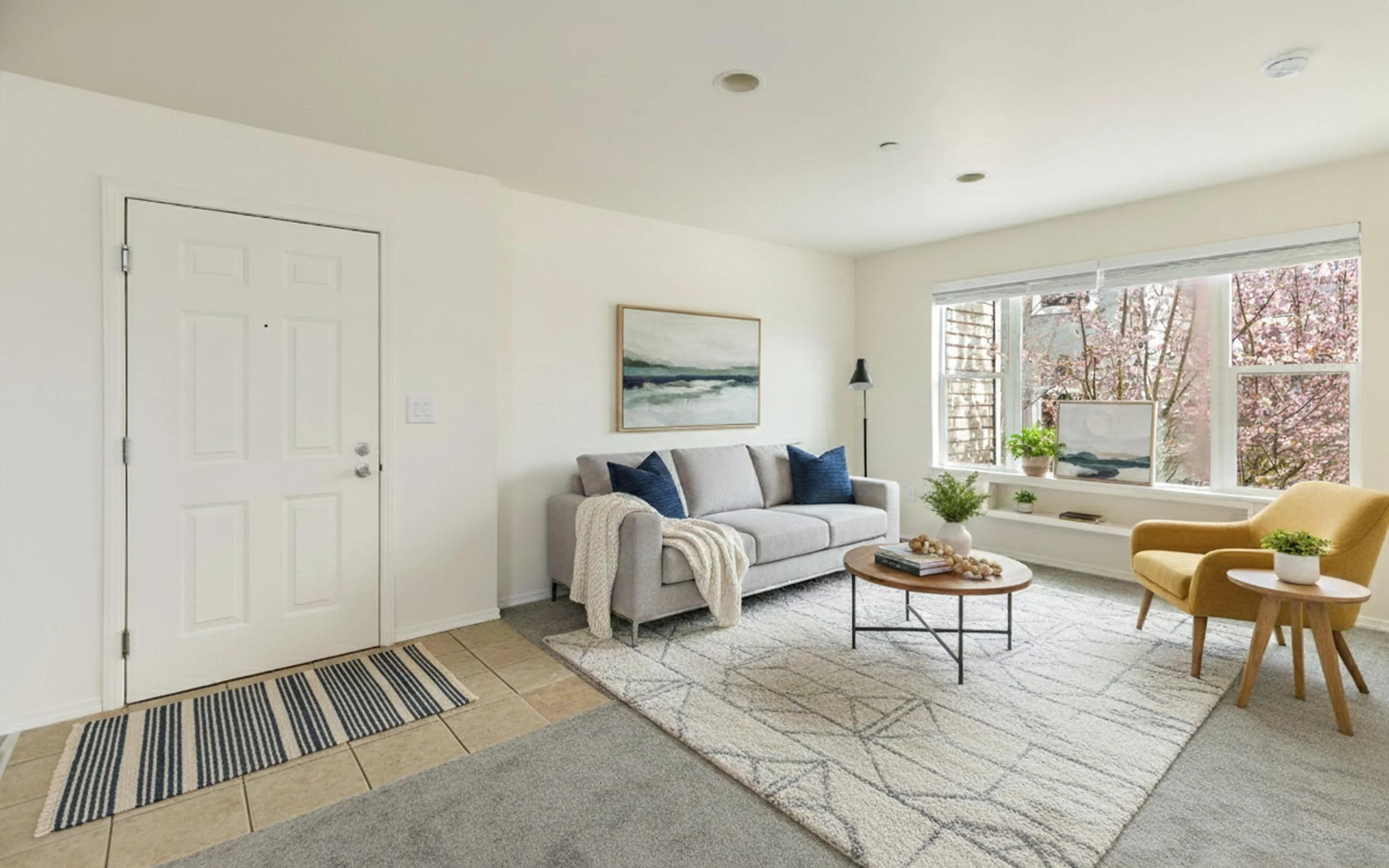 Bright living room with white walls, a white front door, and a large window with pink cherry blossom trees outside. It features a light gray sofa with blue pillows, a yellow armchair, and a round wooden coffee table with plants and books. A white rug