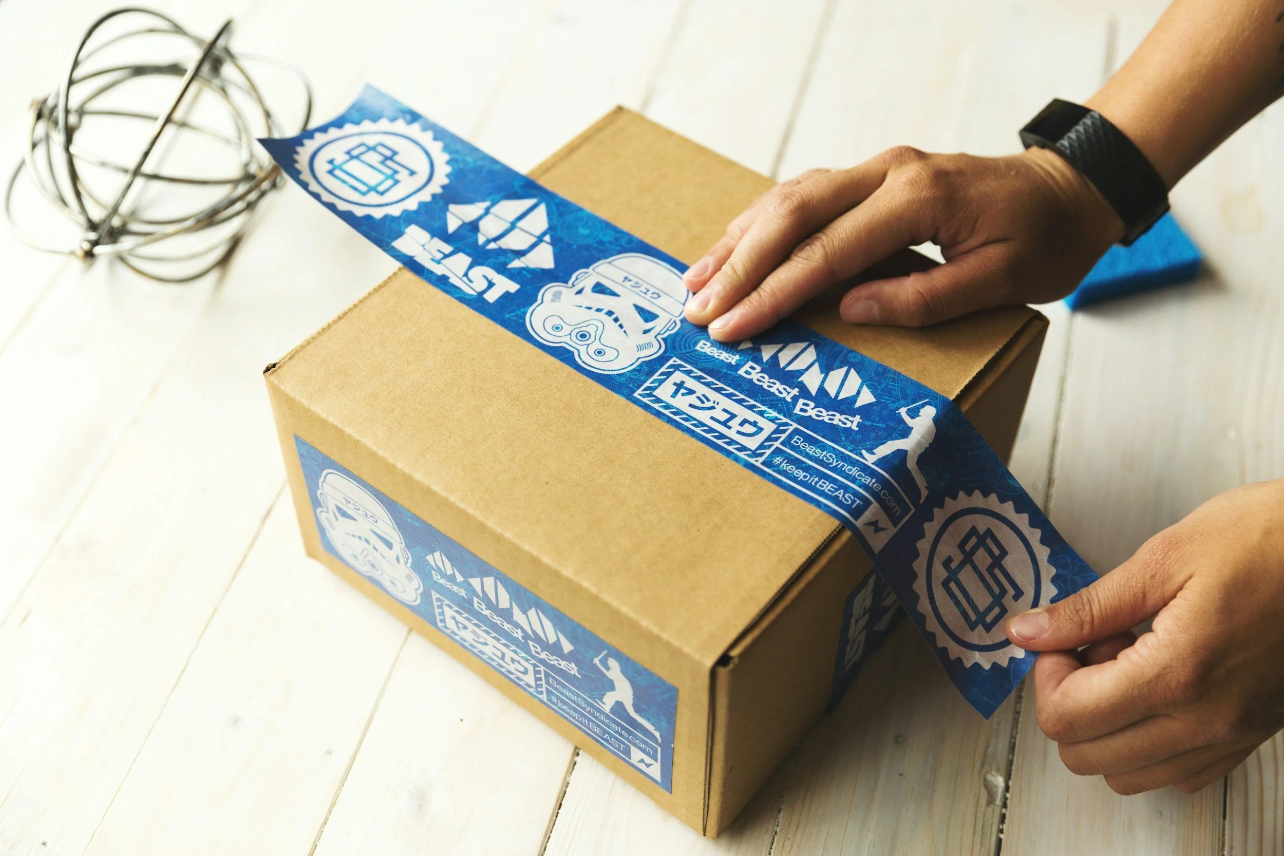 A person sealing a cardboard box with blue tape featuring stickers from Beast Beast, a gym or sports brand, on a white wooden surface.