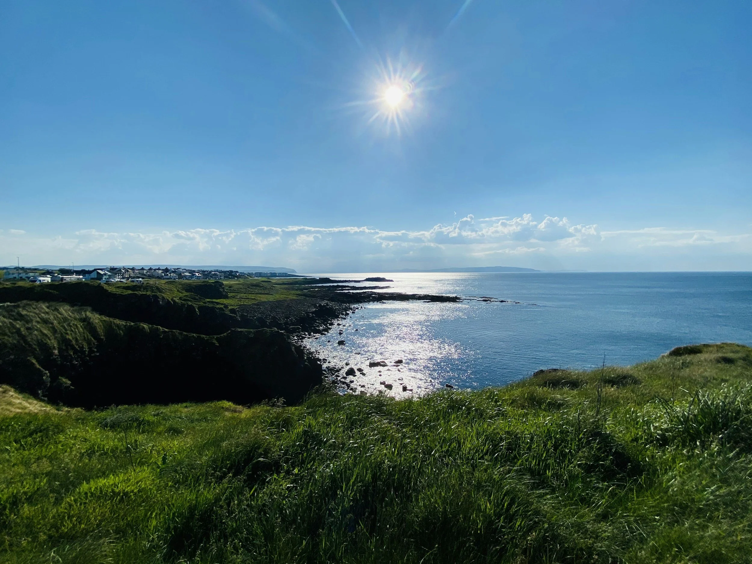 Sunlight reflecting over a calm ocean and green coastline, representing clarity, hope, and peaceful connection on the Holy Facade contact page.