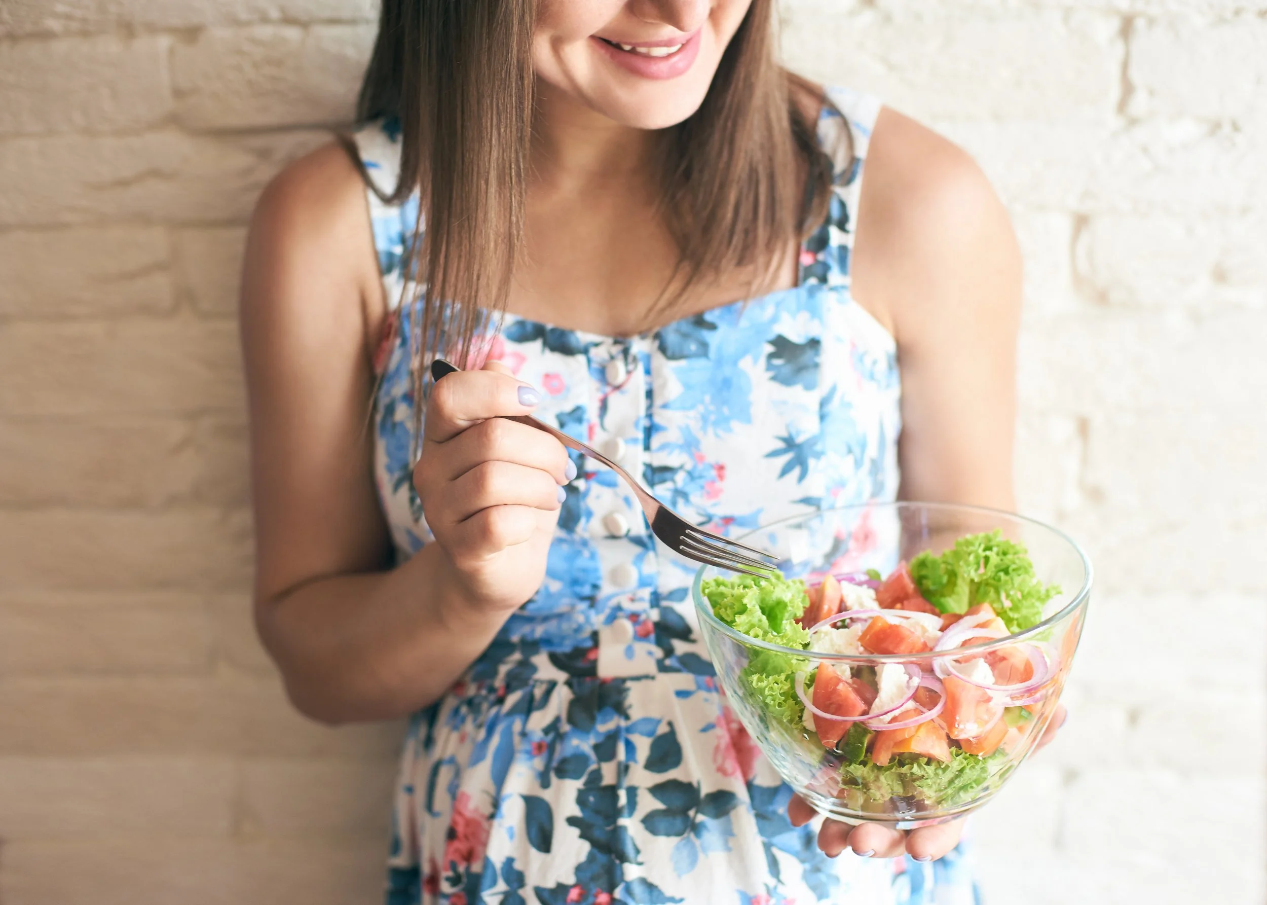 A woman in a floral dress holding a glass bowl of fresh vegetable salad, smiling by a brick wall.