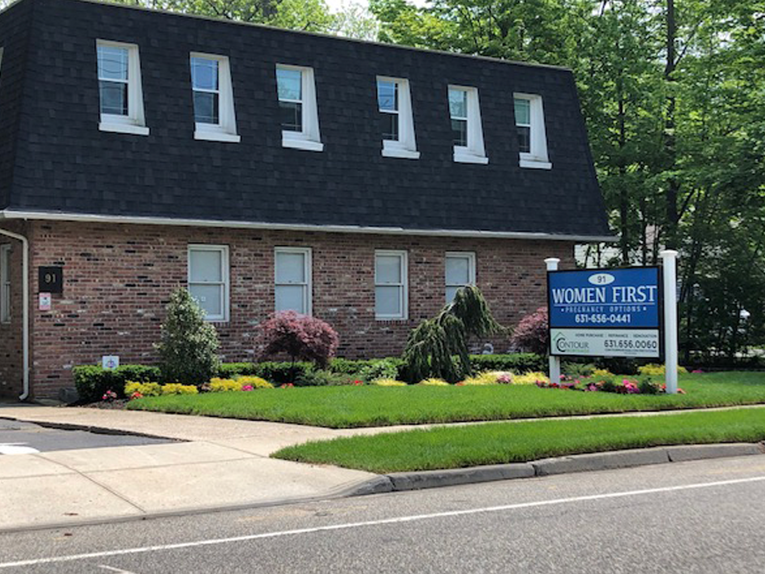 A brick building with a black dormer roof and six small windows. In front, there is a lawn with colorful flowers, a sidewalk, and a street with parking lines. A blue sign reads "WOMEN FIRST" and includes contact information.