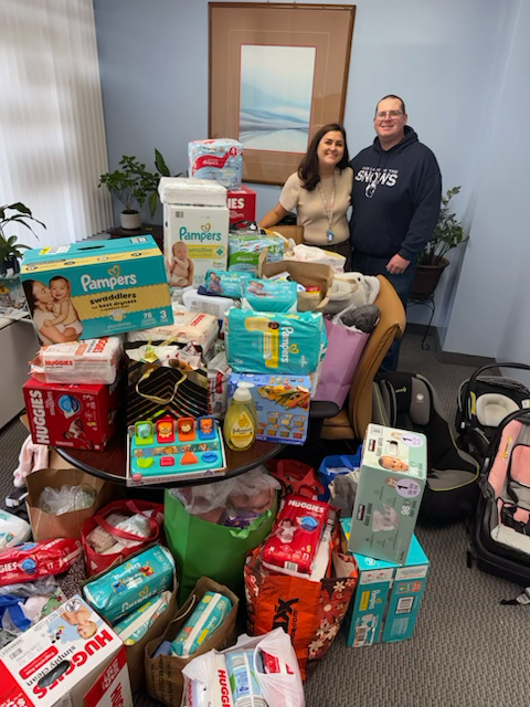 Two people standing behind a large table and piles of baby supplies, including diapers, wipes, and baby formula, in a room with blue walls and framed artwork.