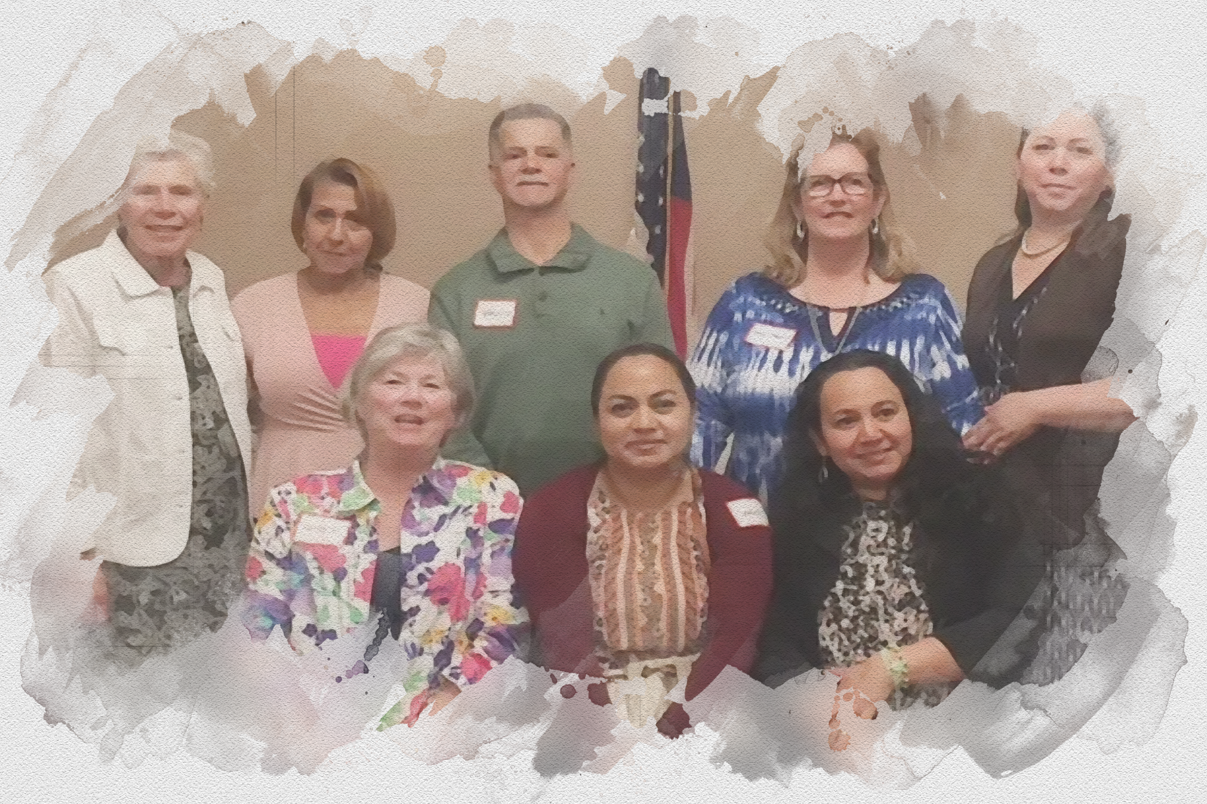 A group of diverse women and one man posing together indoors in front of an American flag, smiling and wearing name tags, at what appears to be a professional or community event.