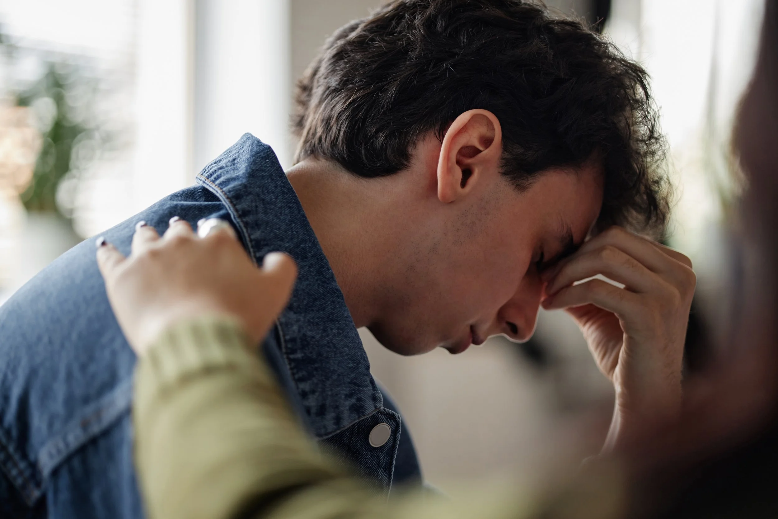 A person with dark, curly hair and light skin holding their head in their hands, appearing stressed or upset. A hand from another person is gently touching their shoulder.