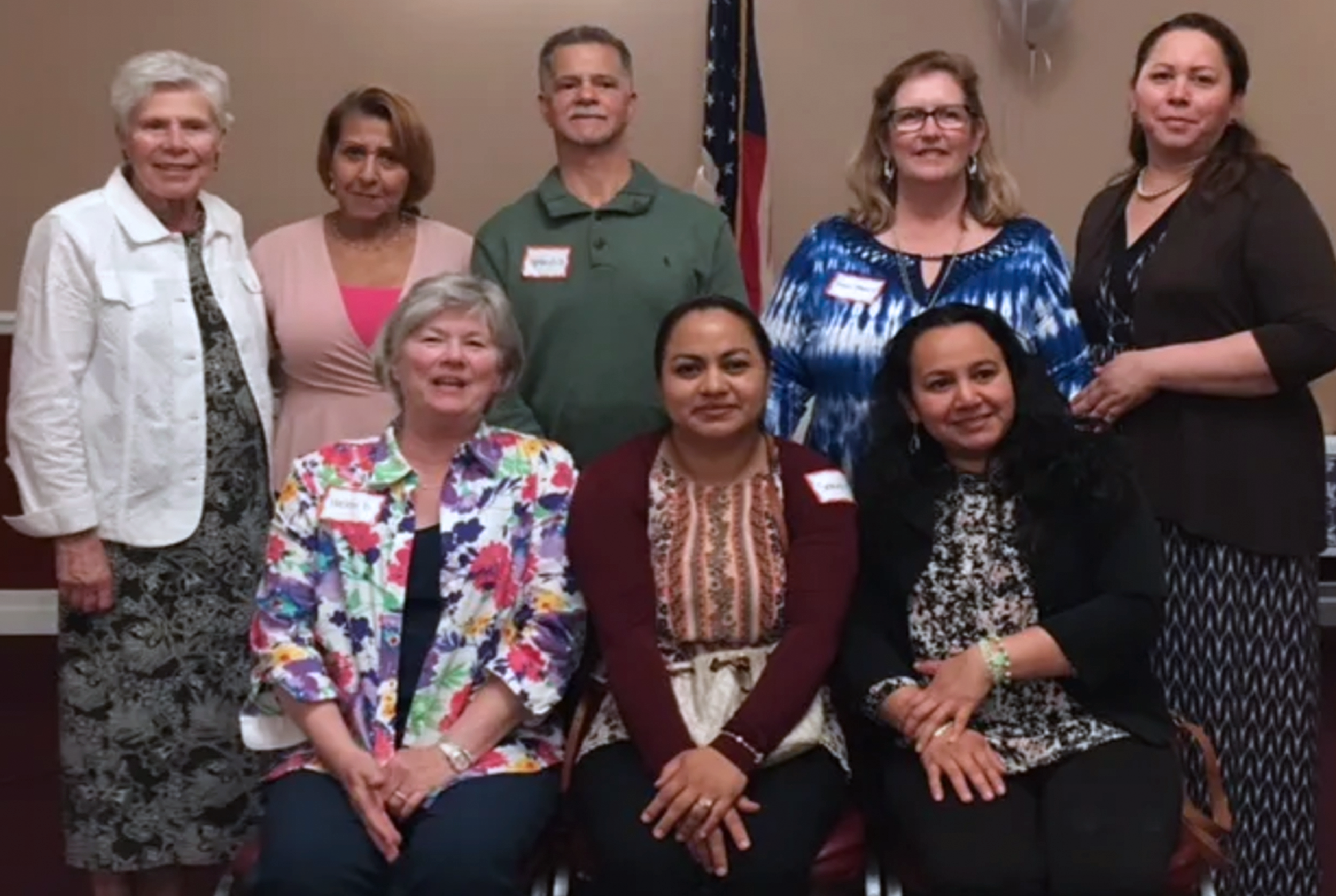 A group of ten women and one man posing for a photo indoors, some wearing name tags, with an American flag in the background.